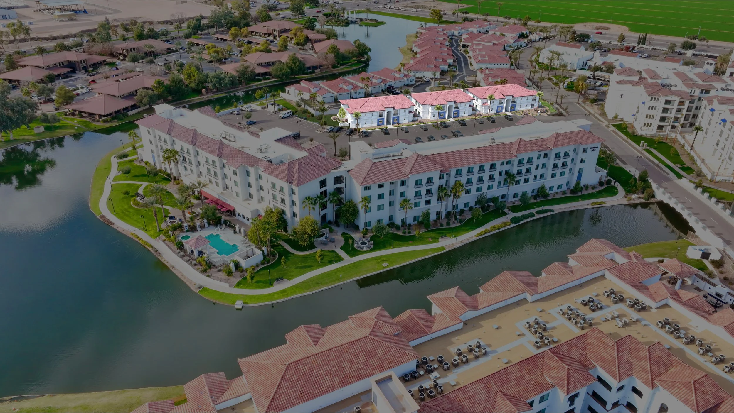 Aerial view of a lakeside apartment complex with red-tiled roofs, a large white building, swimming pool, green lawns, palm trees, water bodies, and surrounding residential buildings.