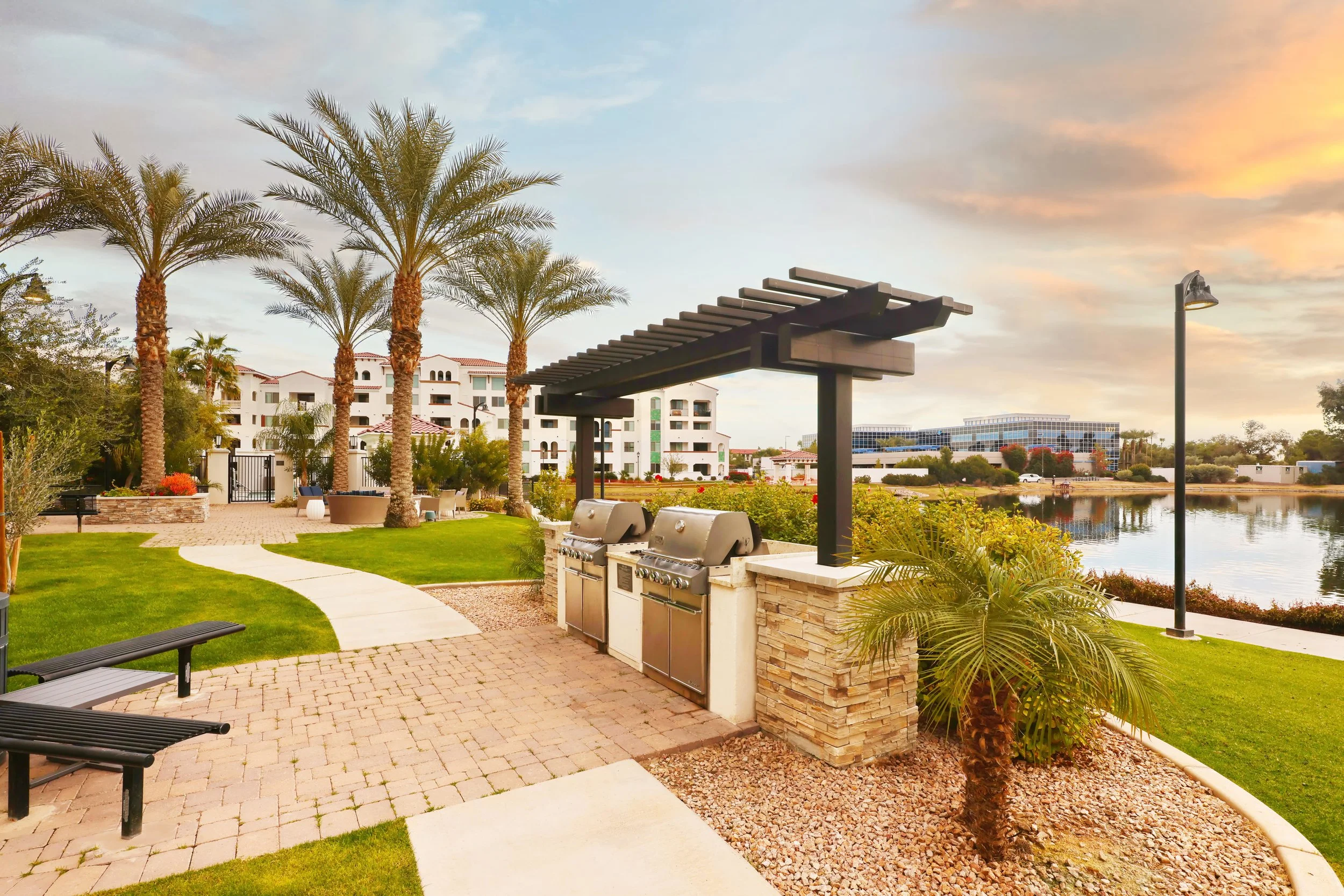 Outdoor park area with palm trees, a brick pathway, walking benches, outdoor grills, a modern trash can, a body of water, and white residential buildings in the background during sunset.