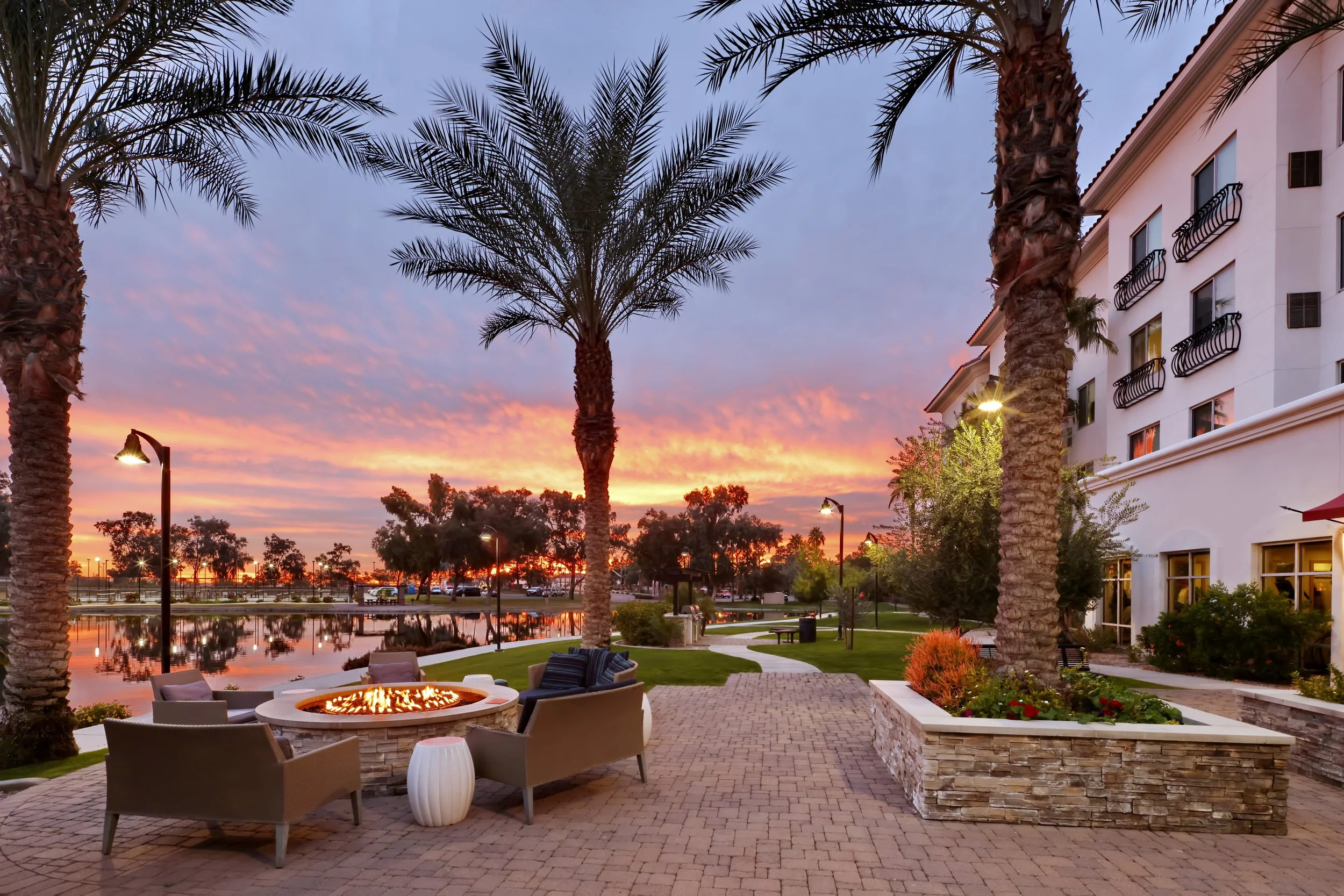 View of an outdoor patio area with a fire pit, surrounded by chairs, palm trees, and a building with balconies, with a sunset sky and trees reflected in a nearby water feature.