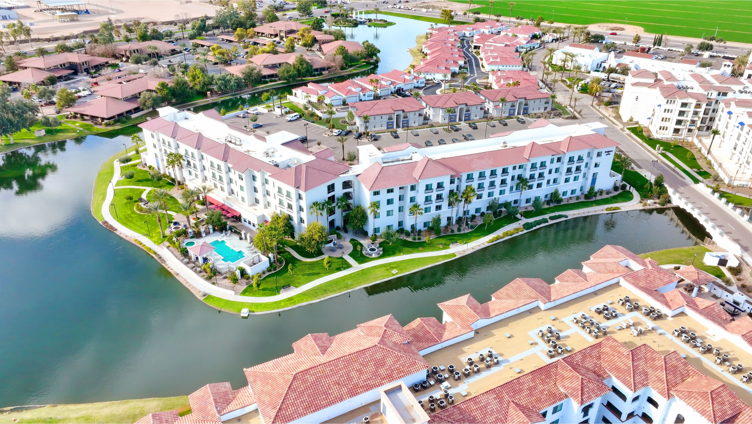 Aerial view of a residential complex with white buildings, red-tiled roofs, water features, and green lawns, with parking areas and surrounding roads.