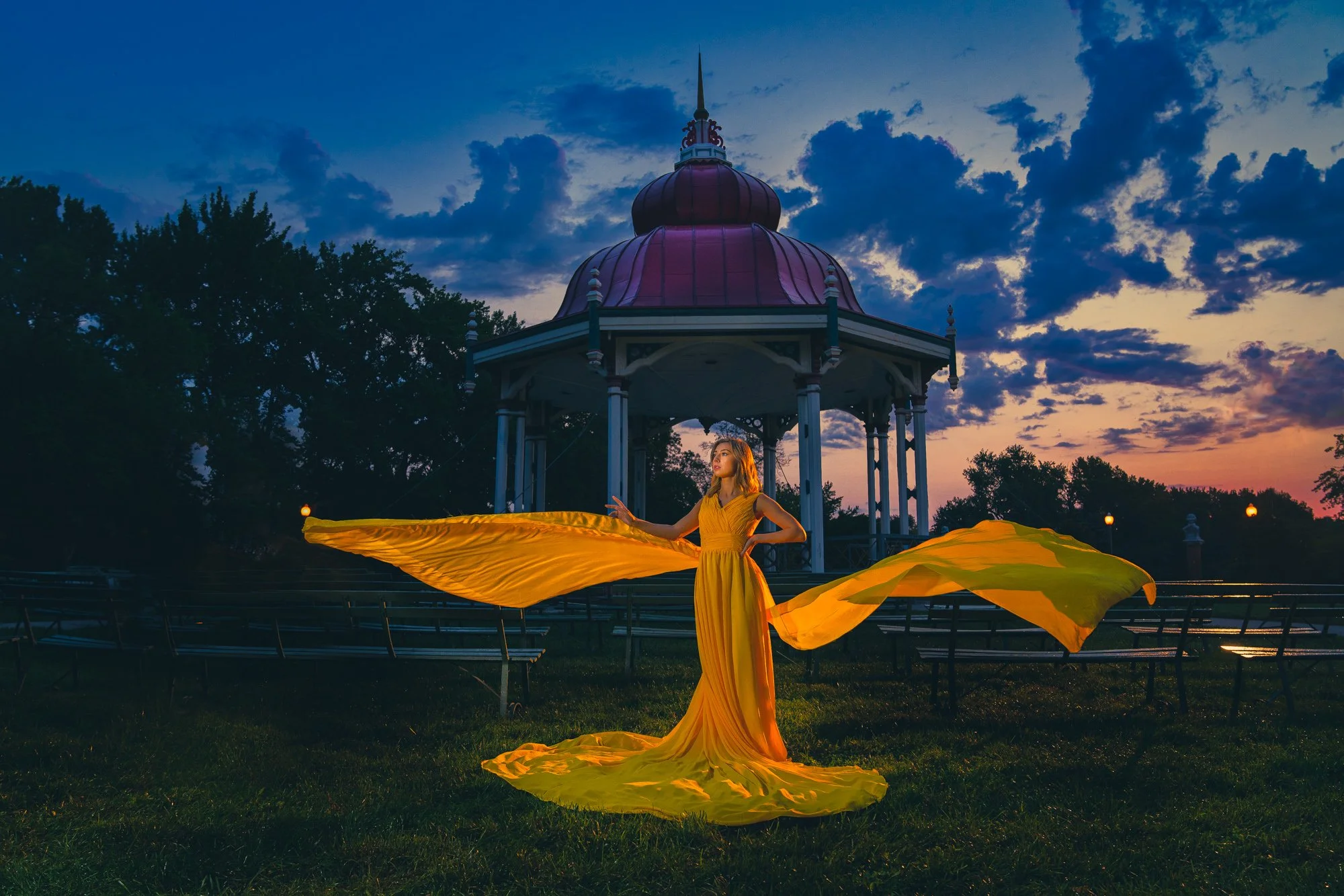 High school senior in a flowing yellow gown posing near a gazebo at sunrise.