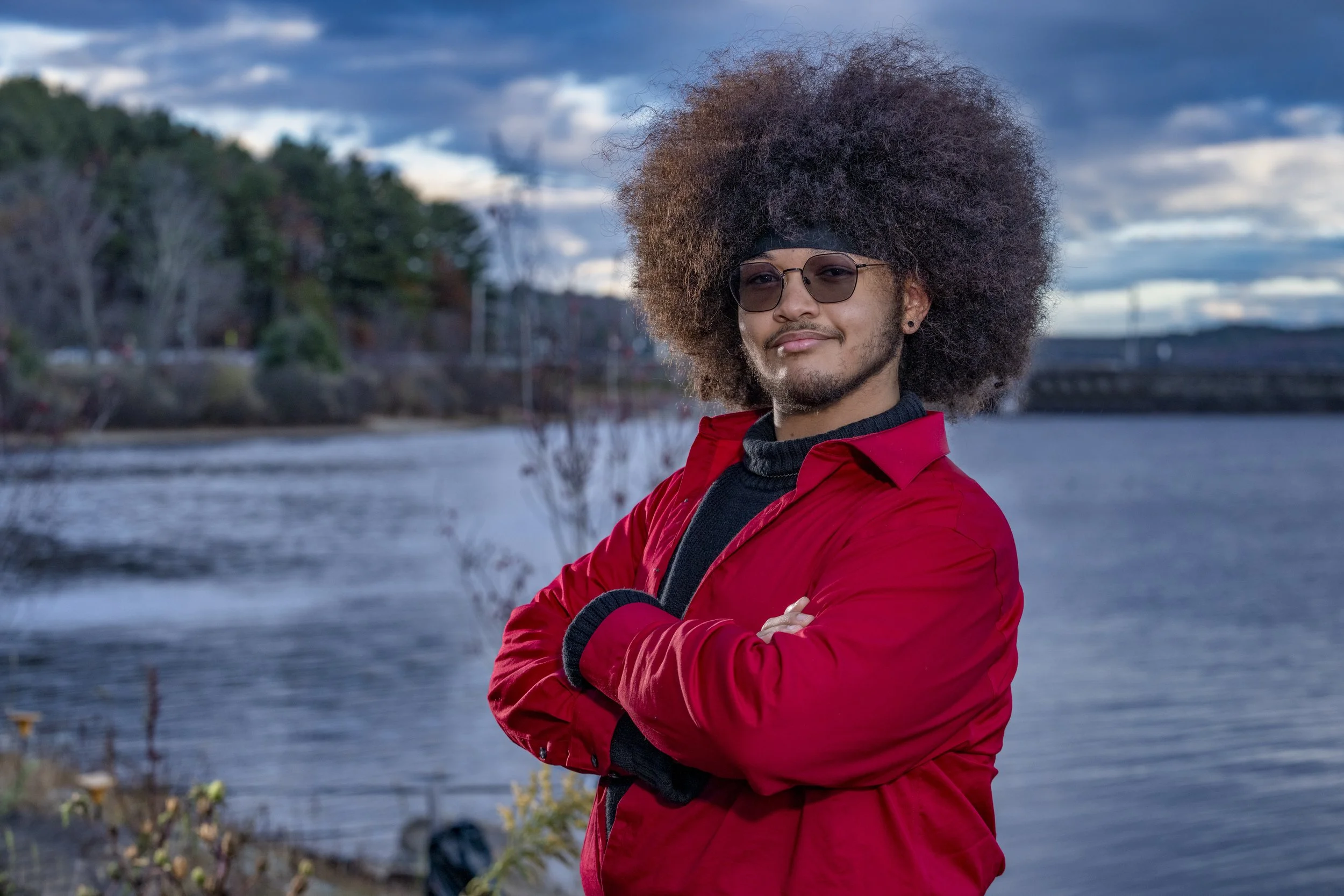 Male high school senior wearing a red jacket and fur hat beside the water in Southern New Hampshire