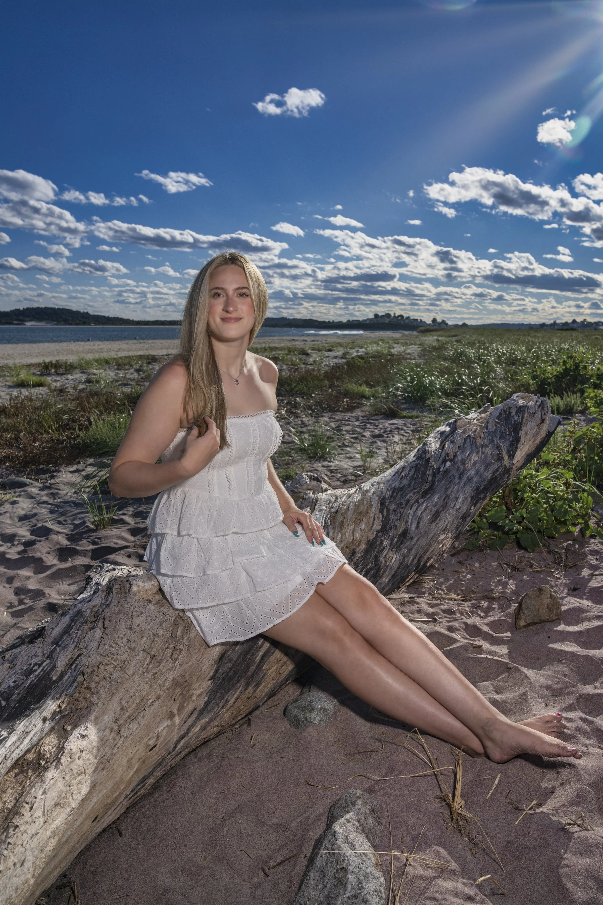 High school senior in a white dress seated on driftwood during a beach portrait session in Northern Massachusetts.