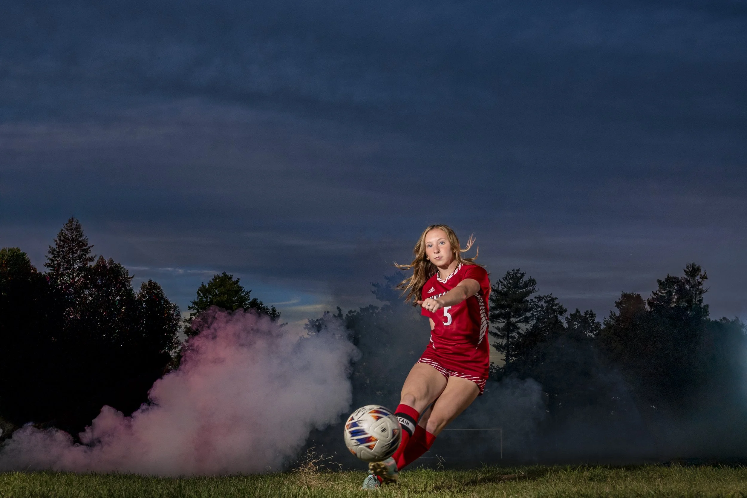 Female high school soccer player kicking a ball through pink and blue smoke during a creative senior sports portrait