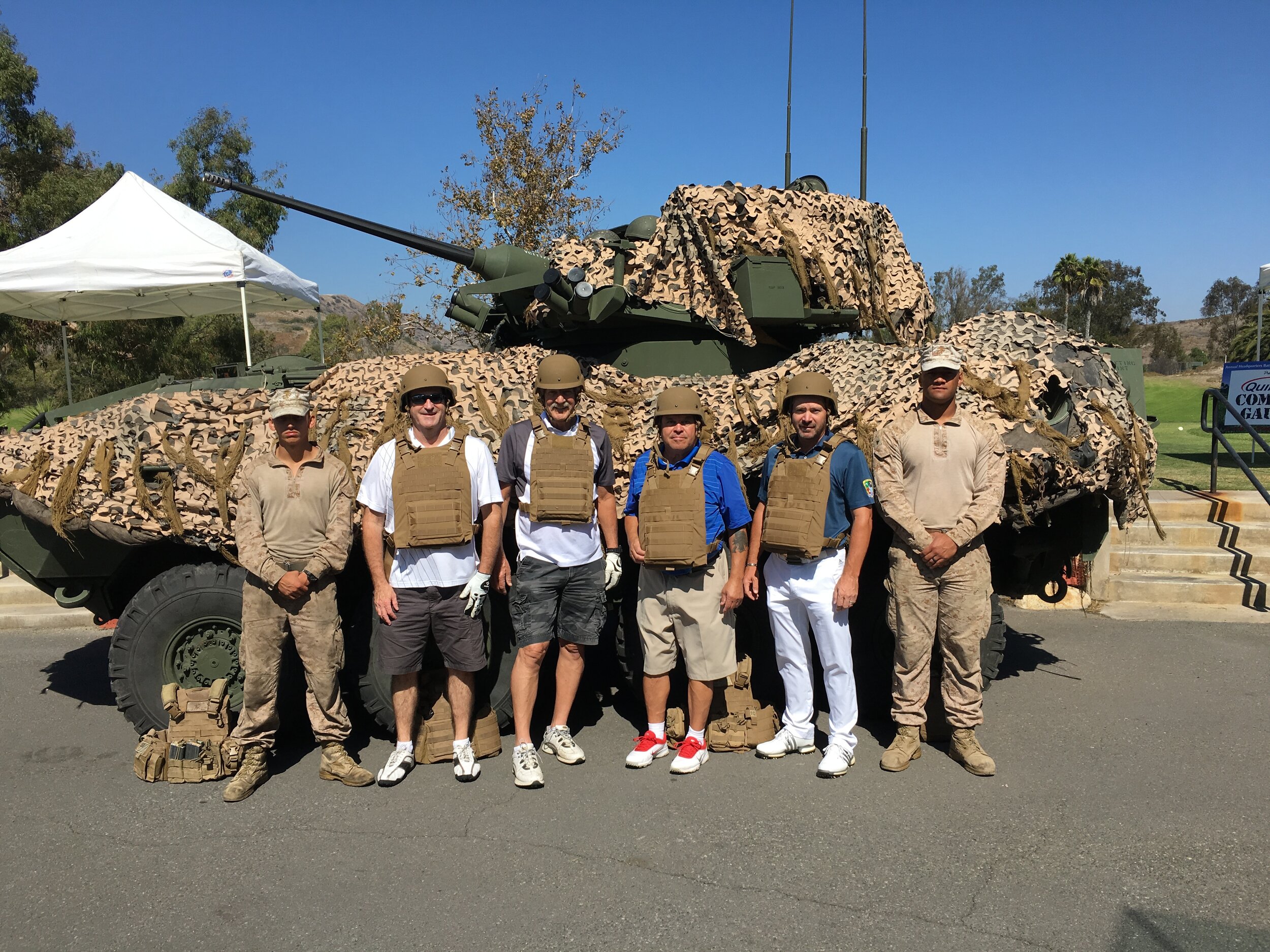 Group of six men in military gear and casual clothing standing in front of a military tank with camouflage netting on it. They are wearing helmets and tactical vests, with some smiling at the camera. The background includes trees, a white tent, and s