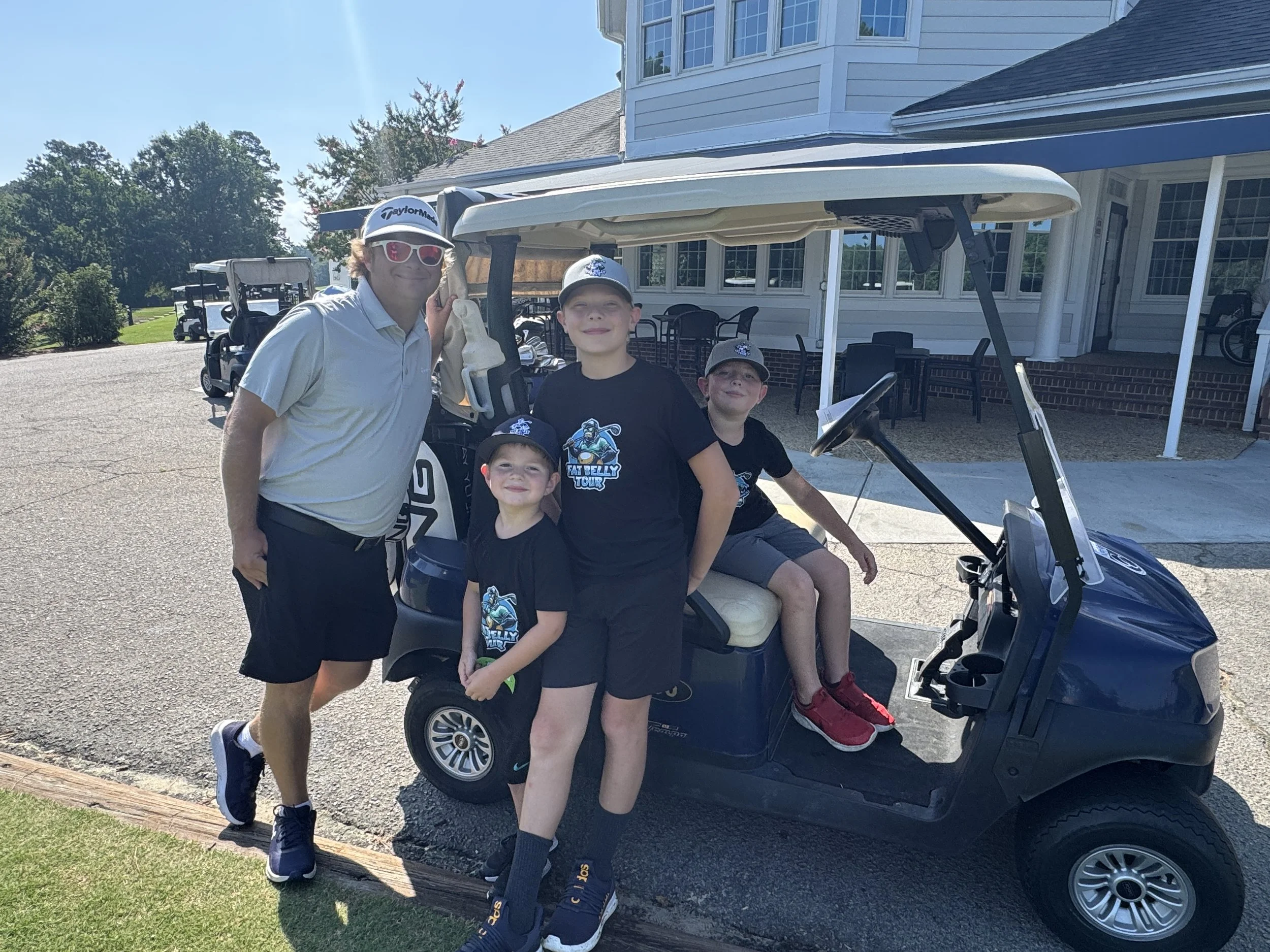 A man and three boys posing next to a blue golf cart outside a large white building with a porch. The boys are wearing black t-shirts with a hockey logo, and the man is wearing a white polo shirt, black shorts, sunglasses, and a cap. The boy sitting in the golf cart is wearing red shoes, and the other two boys are standing beside the cart.