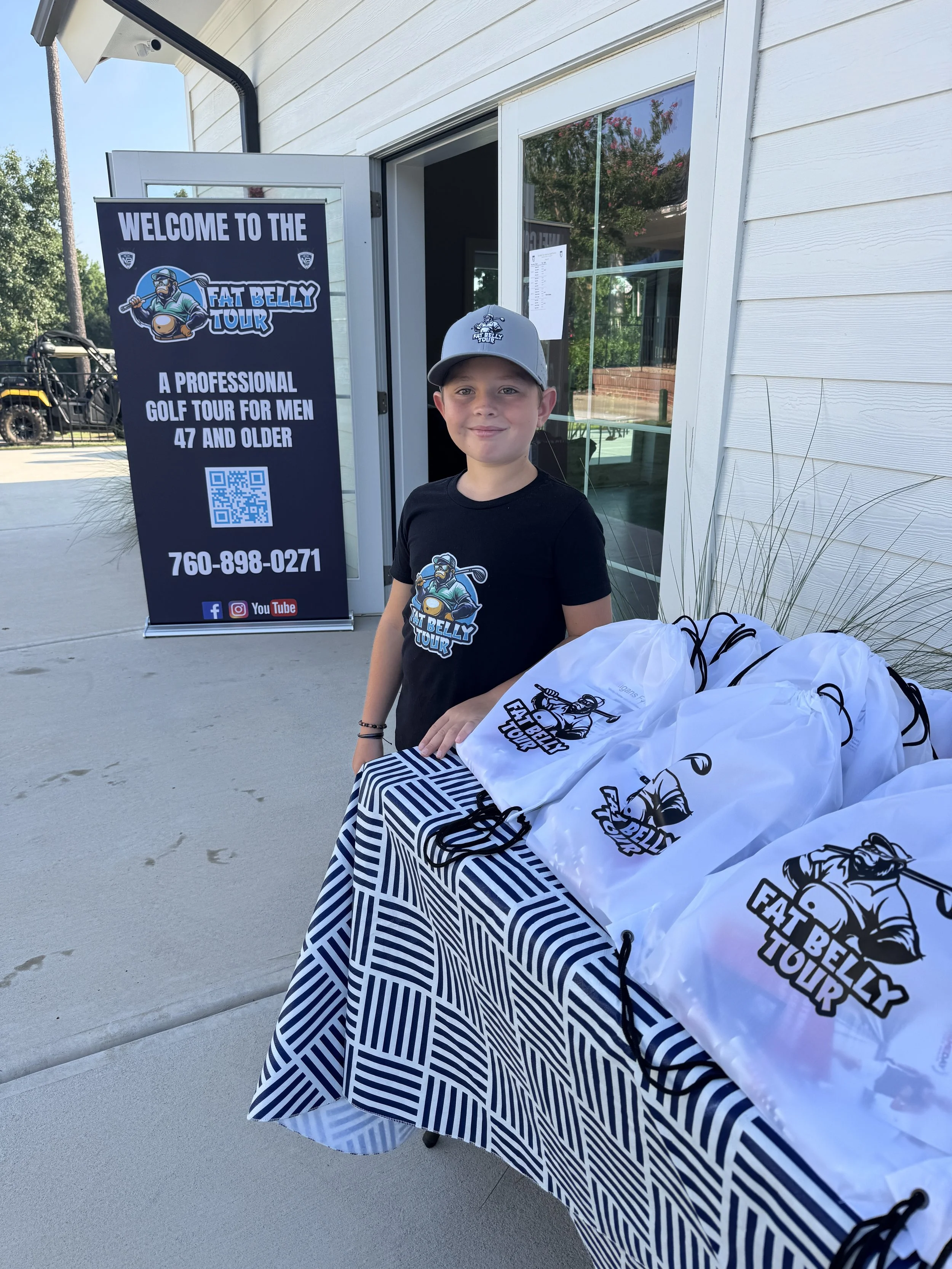 Young boy standing beside a table with white drawstring bags, wearing a black T-shirt and gray cap, at an outdoor event named Fat Belly Tour for men 47 and older, with a sign in the background displaying the event logo, phone number, and social media icons.