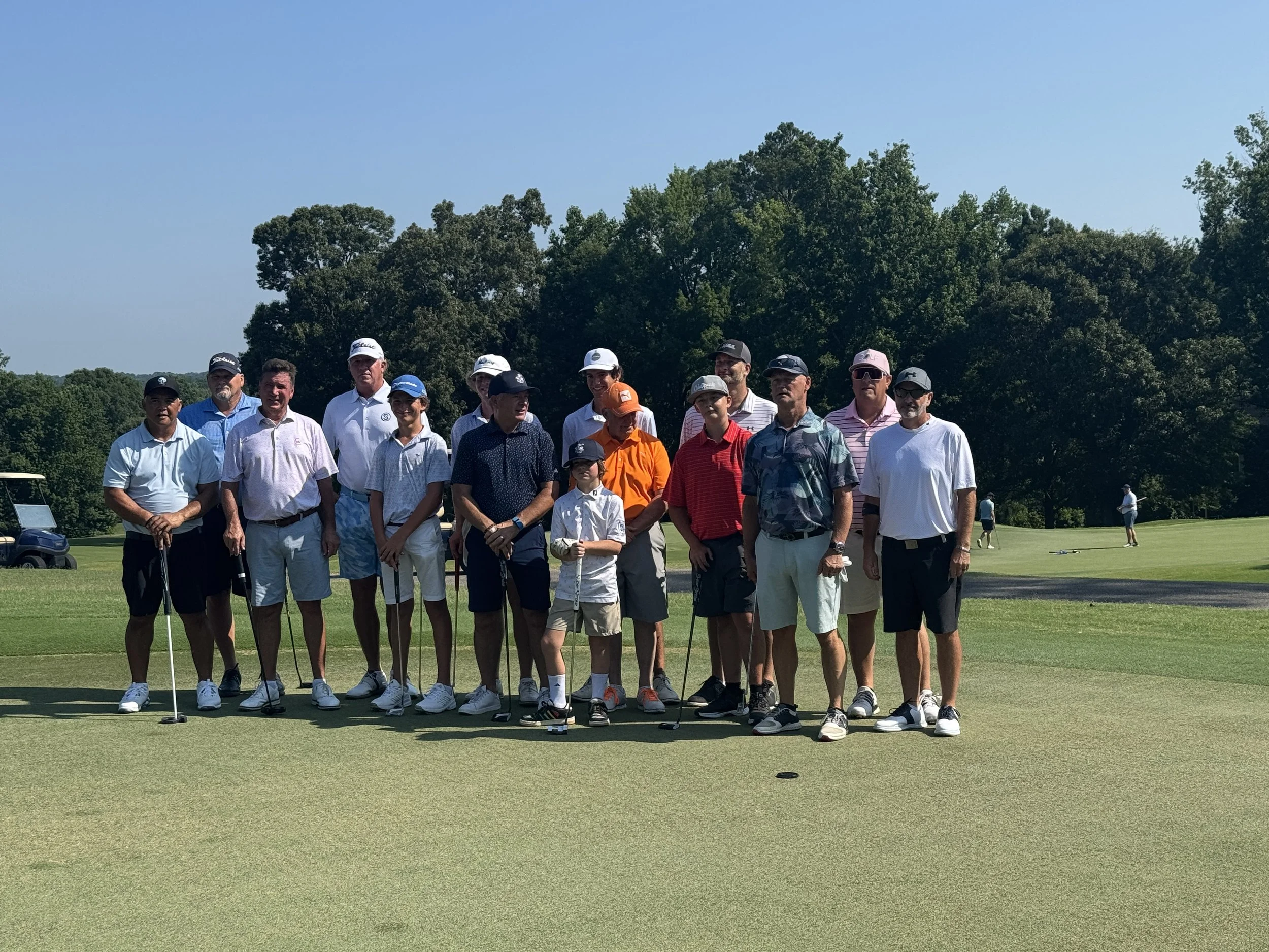 Group of people on a golf course, standing on the green with golf clubs, with trees in the background.