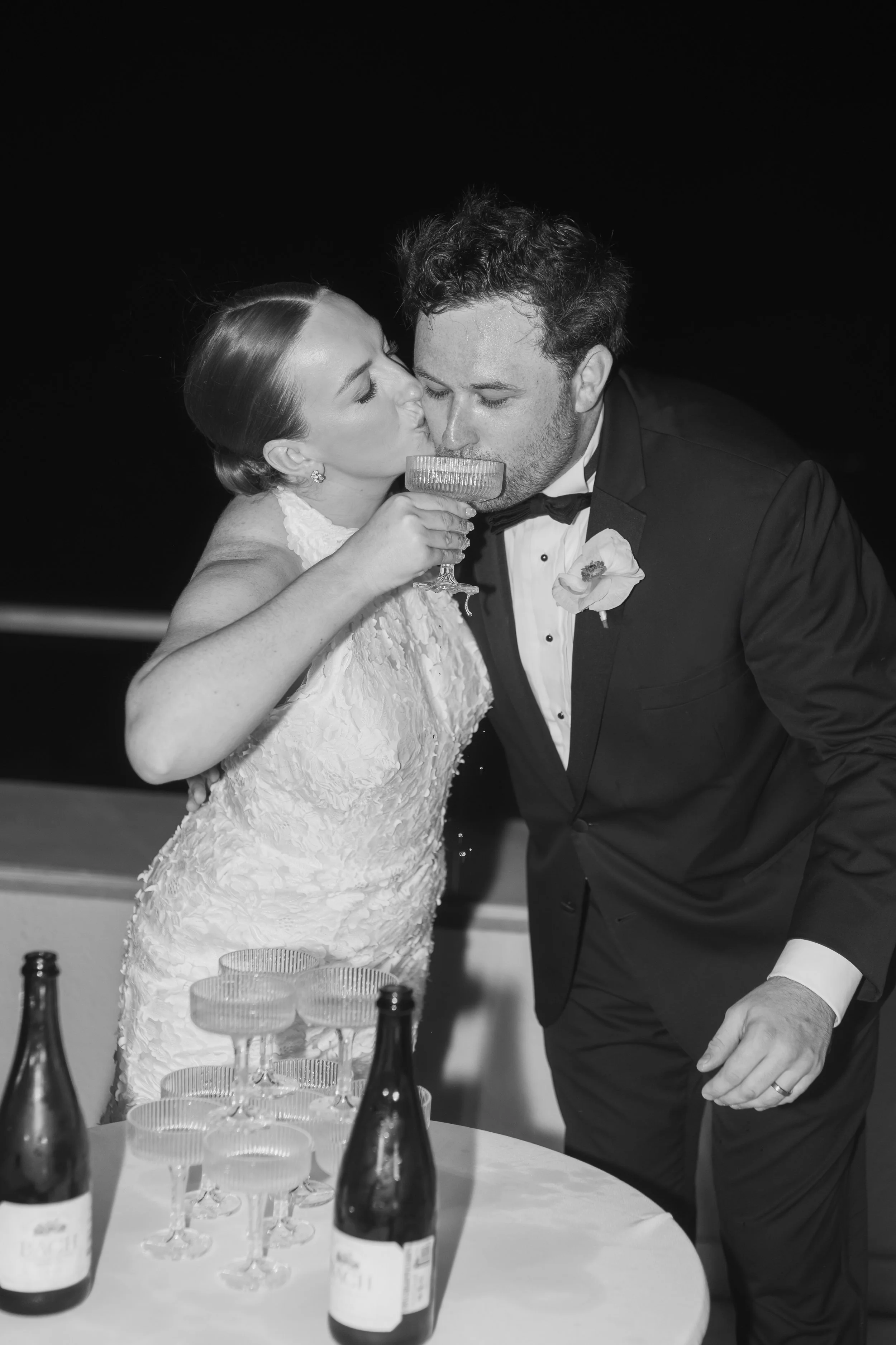 A bride and groom sharing a toast at their wedding reception, with a tiered cake and champagne bottles on the table in front of them.