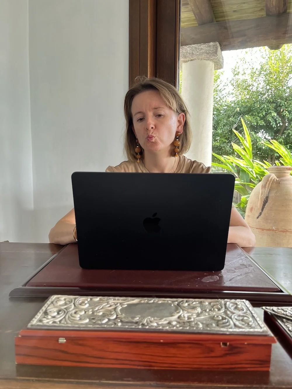A woman sitting at a wooden table, looking at her open black MacBook laptop with a curious or surprised expression. Behind her, a window shows green foliage and sunlight.