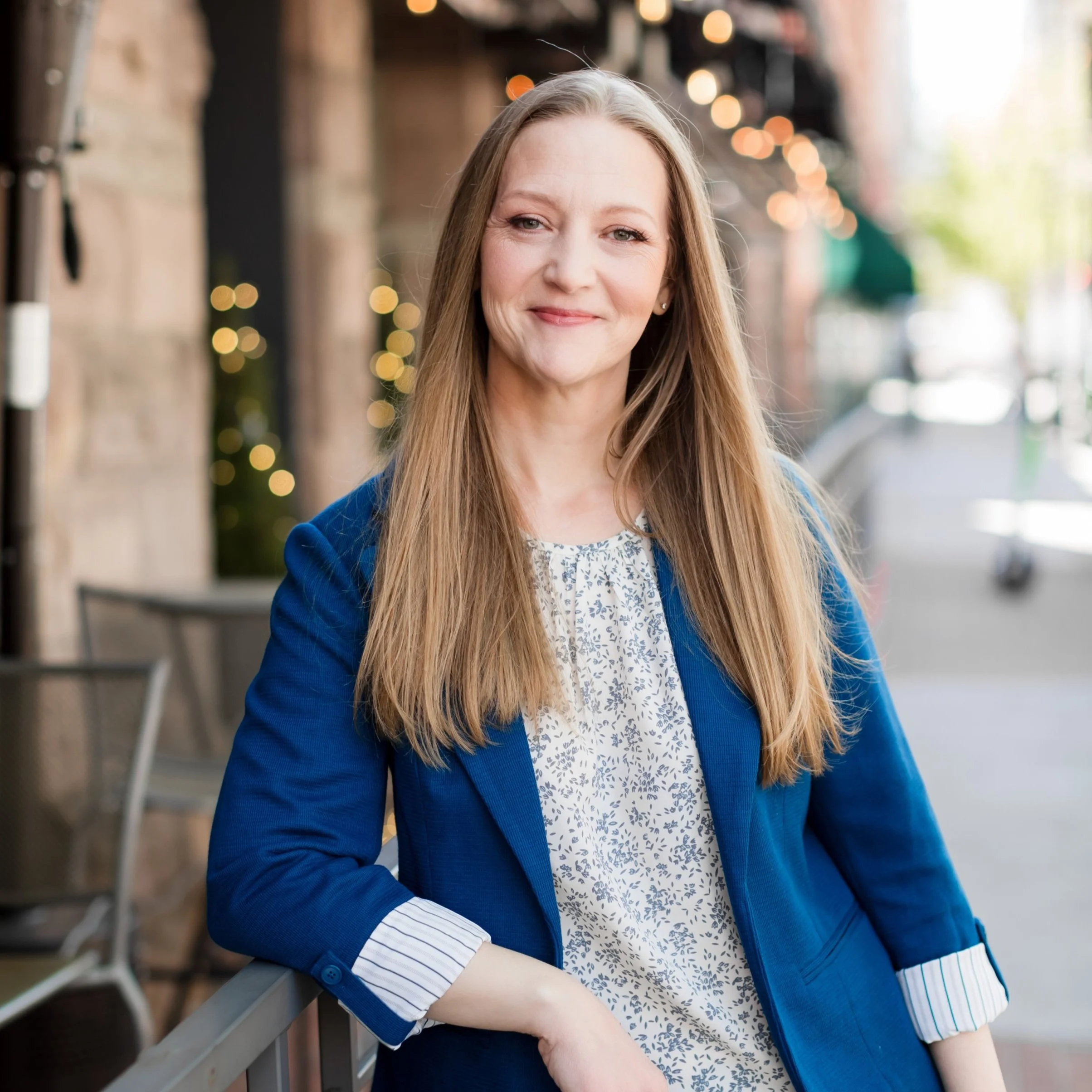 A woman with long blonde hair smiling outside on a city sidewalk, wearing a blue blazer and a white patterned blouse, with blurred storefronts and string lights in the background.