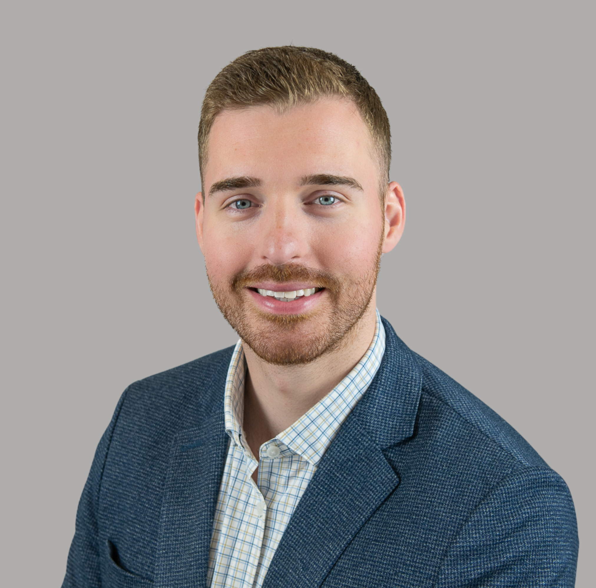 Headshot of a young man with light brown hair, blue eyes, and a beard, wearing a blue blazer and a light checkered shirt, smiling against a gray background.