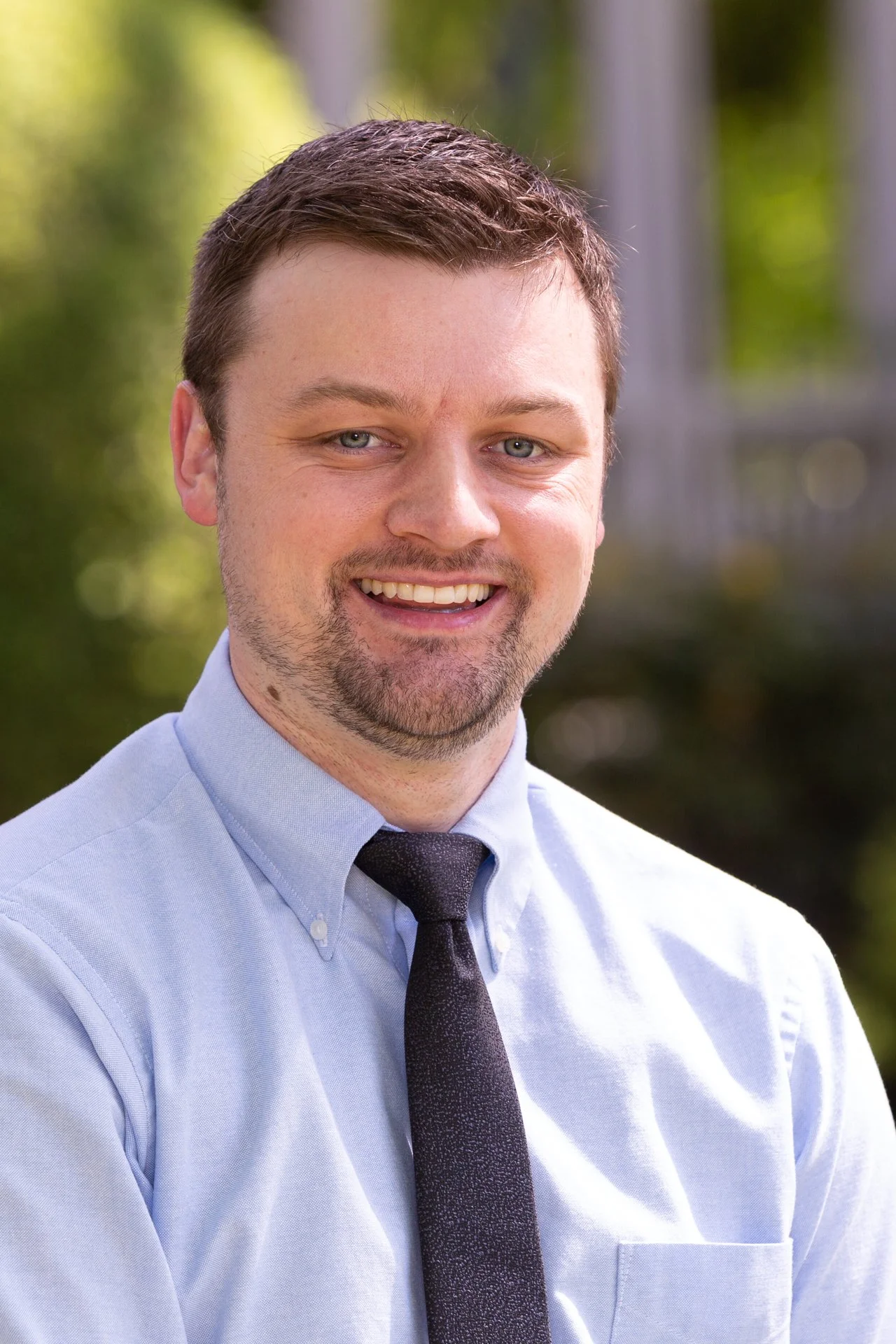 A smiling man in a light blue dress shirt and black tie outdoors with blurred greenery in the background.