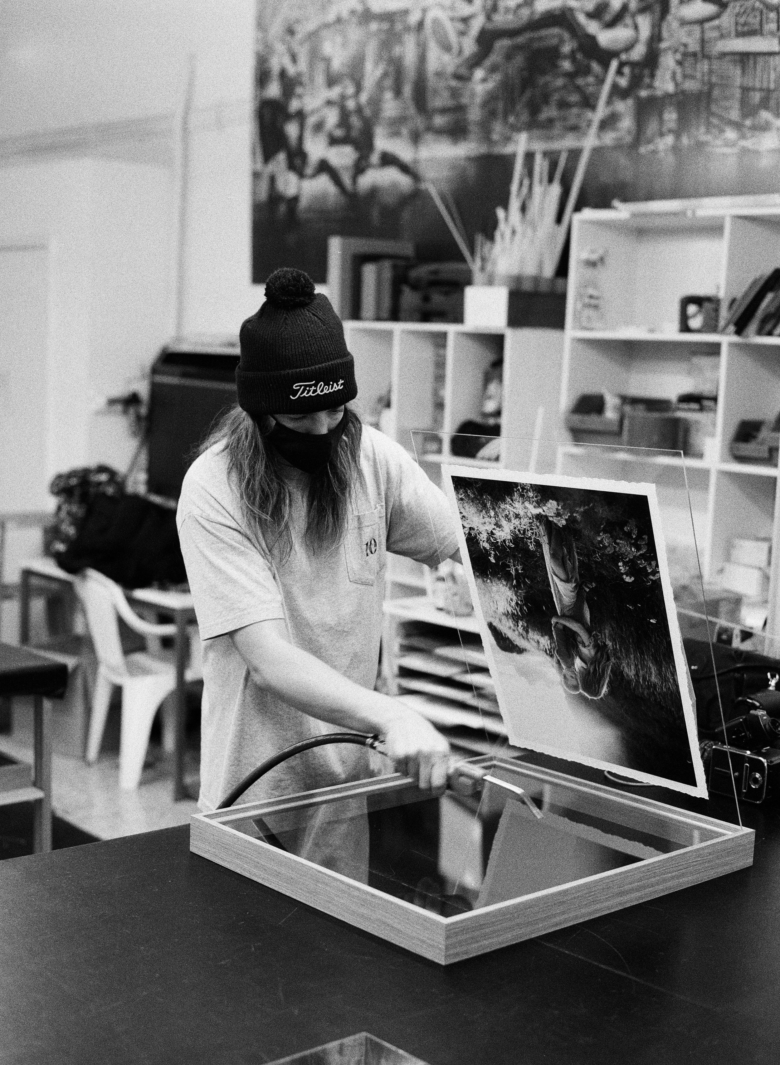 A woman wearing a beanie, mask, and T-shirt is working with a heat press machine on a photograph of a woman with curly hair, displayed inside a glass case in a room with shelves and artwork.