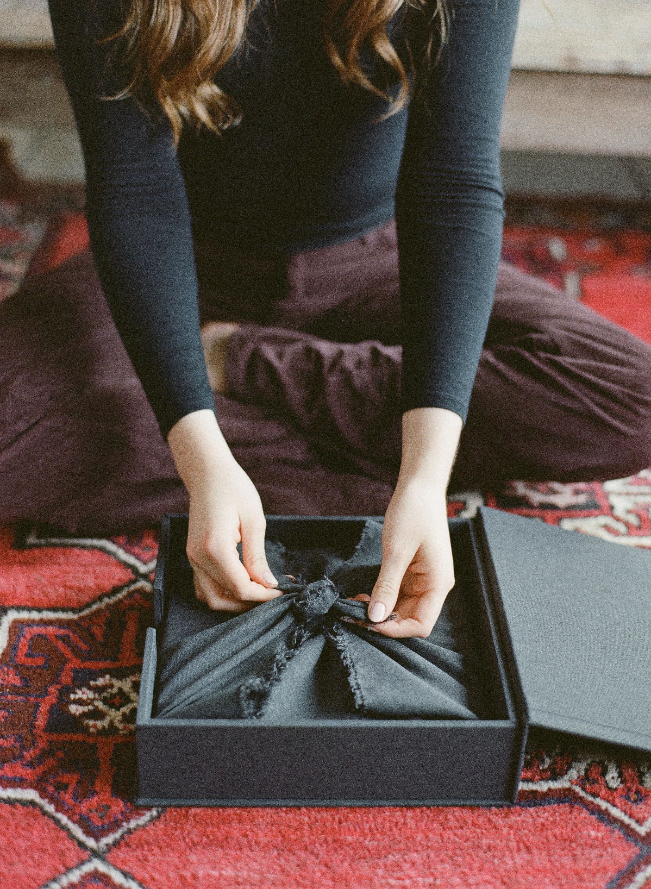 Person sitting on a patterned red and black rug, wearing a black long sleeve shirt and burgundy pants, opening a black box to reveal folded black fabric inside.