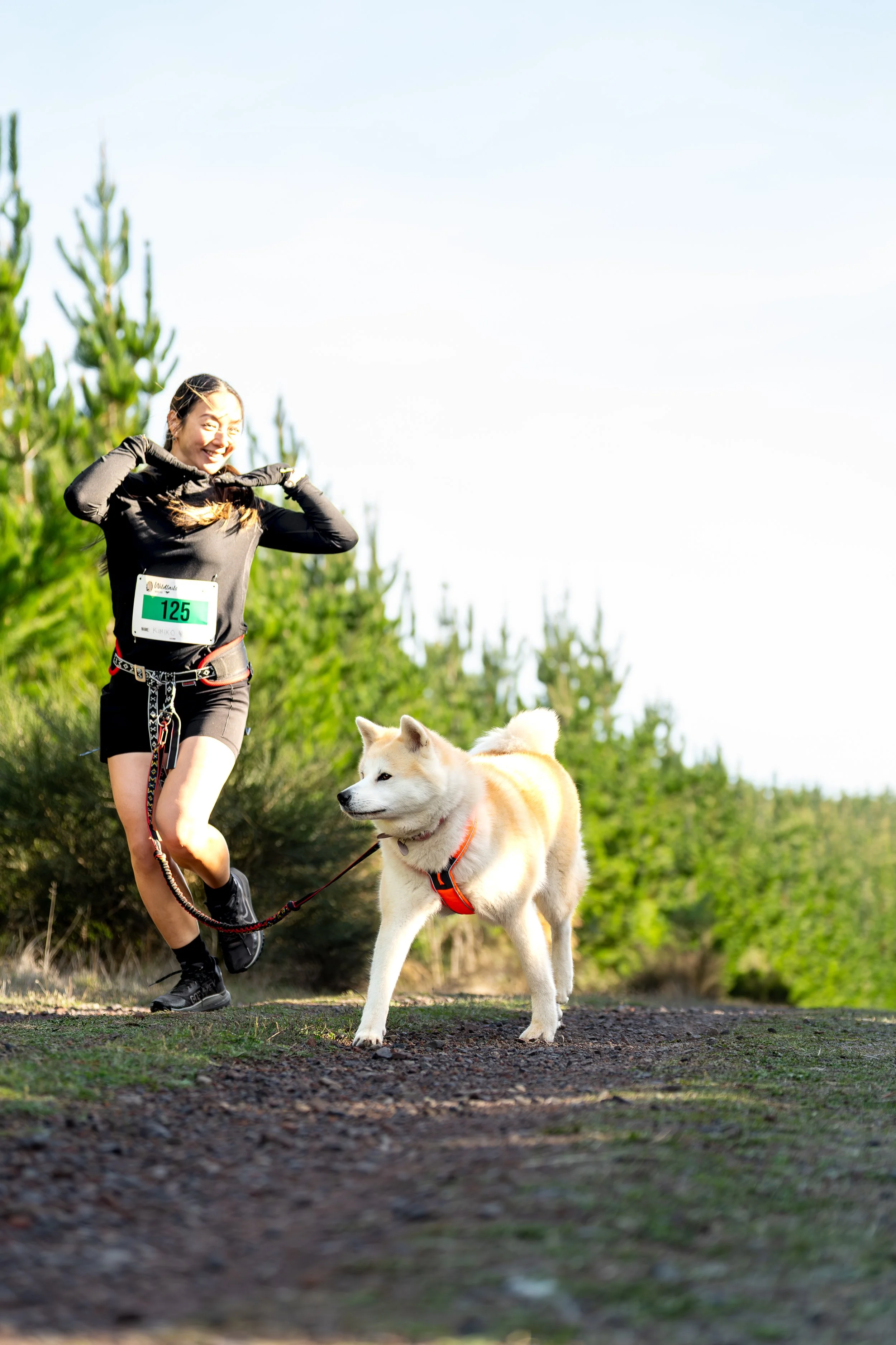 A woman running with her dog on a trail in a forested area, wearing a bib with the number 125.