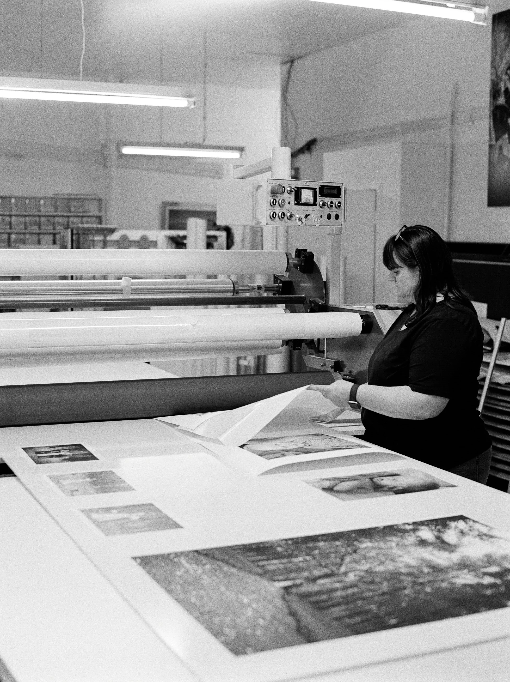 A woman operating a large-format printer in a print shop, with printed photographs laid out on the table.