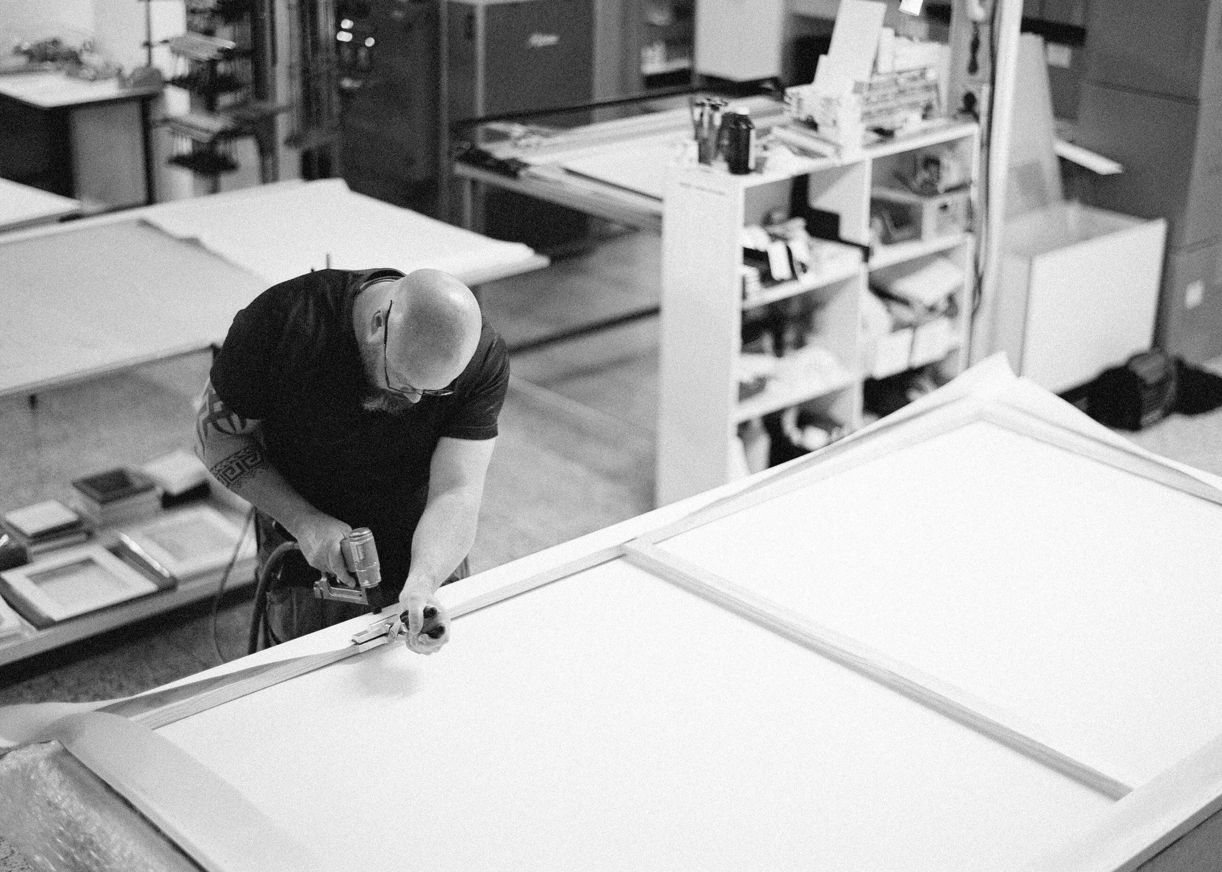 A man working on a large piece of furniture or artwork in a workshop, using a power drill, surrounded by shelves and work tables.