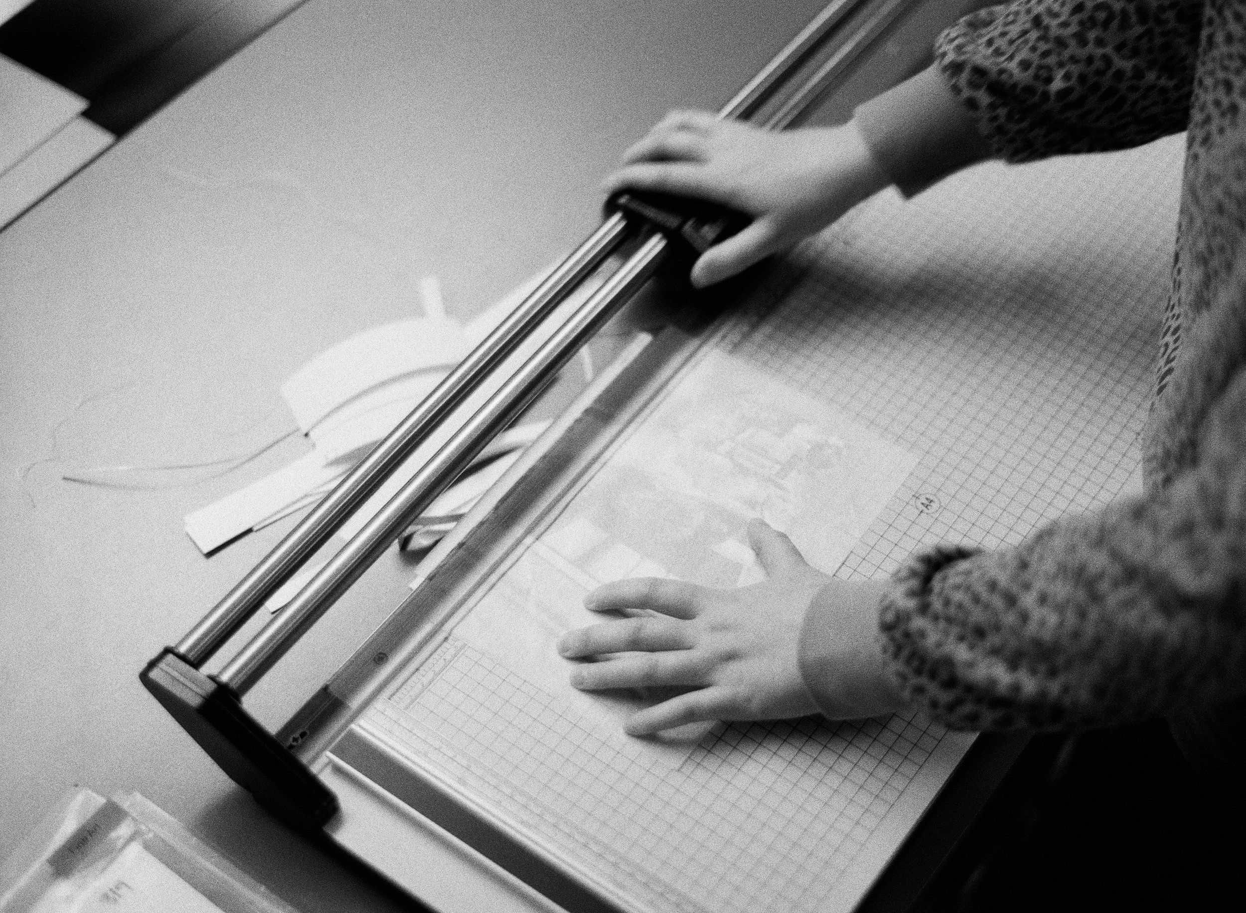 Child in patterned sweater using a paper cutter on a grid-lined cutting mat with paper strips nearby.