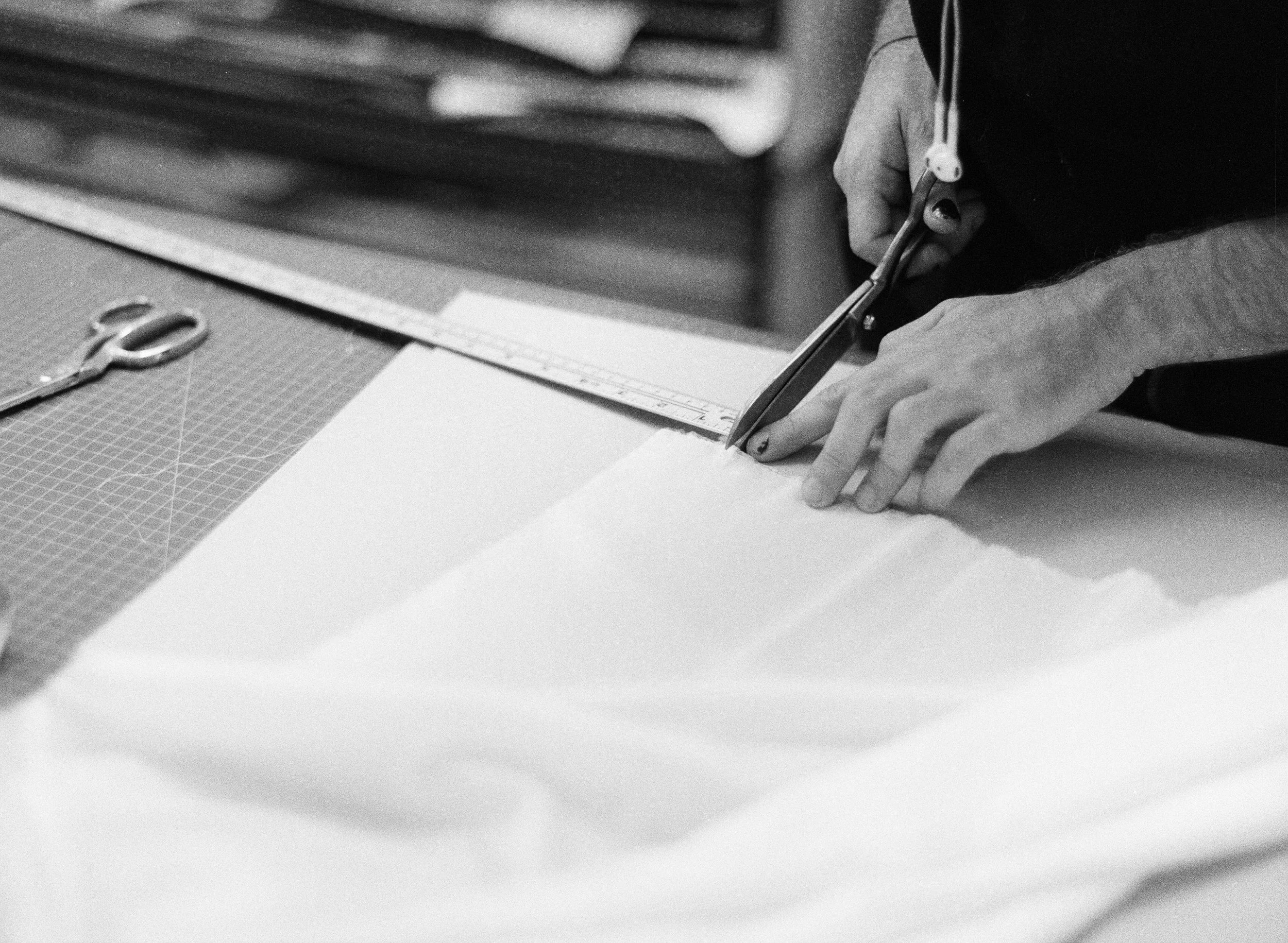 A person is cutting white fabric with scissors on a worktable, using a straight edge guide. There are scissors and a cutting mat on the table.
