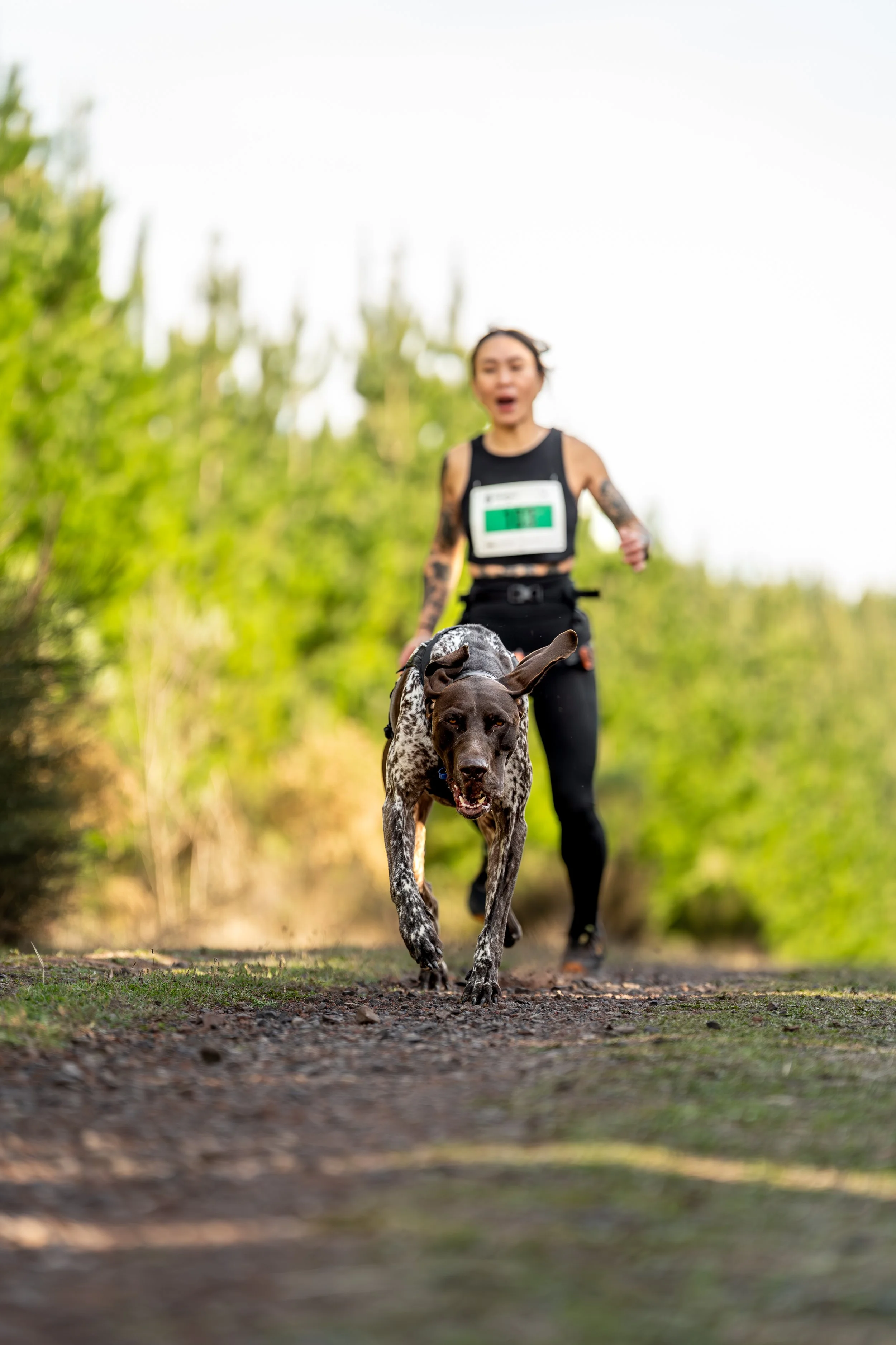 A woman running with her dog on a dirt trail through a forested area.