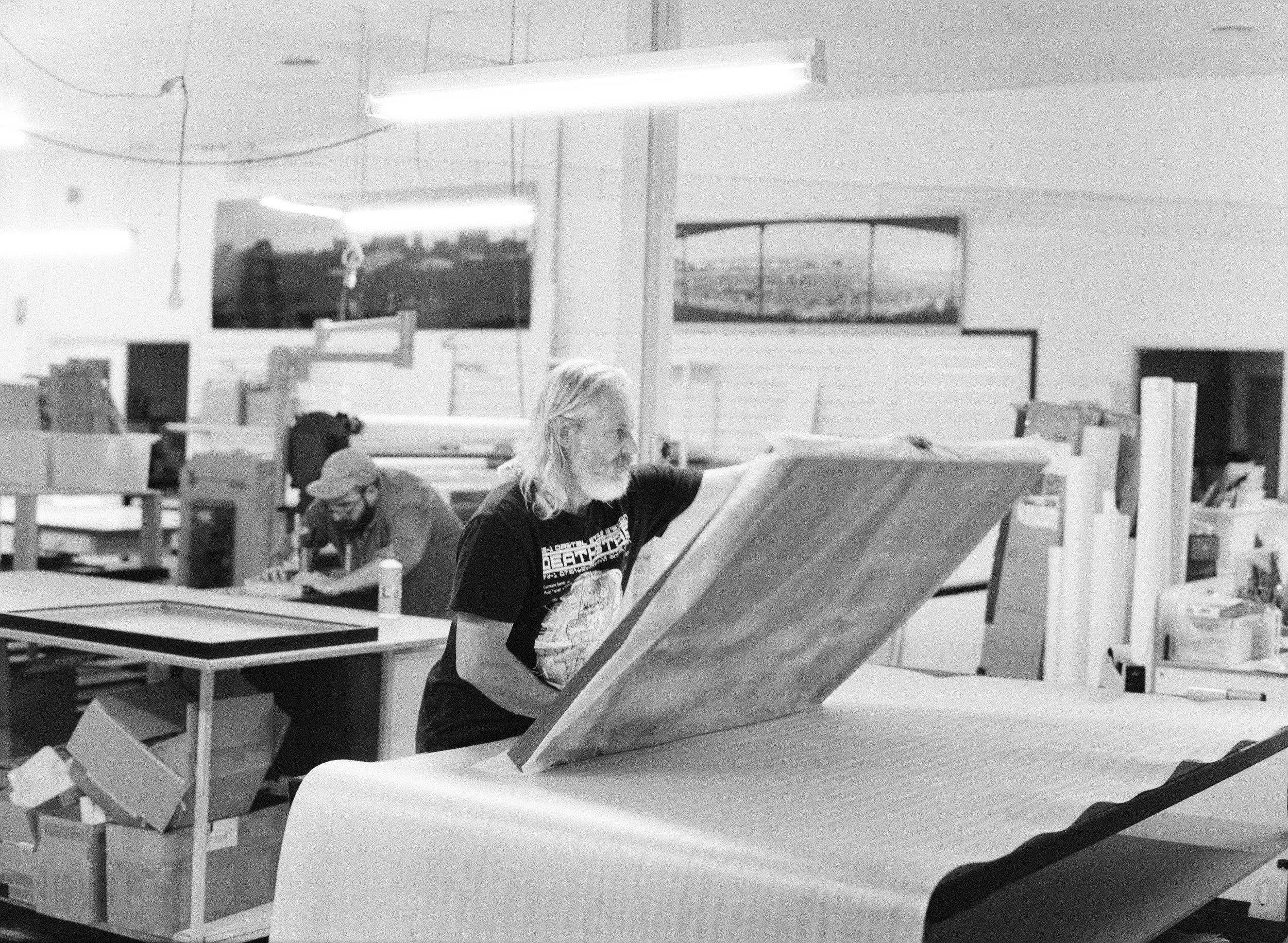 A man with long hair and a beard working on a large piece of wood in a workshop, with another person in the background.