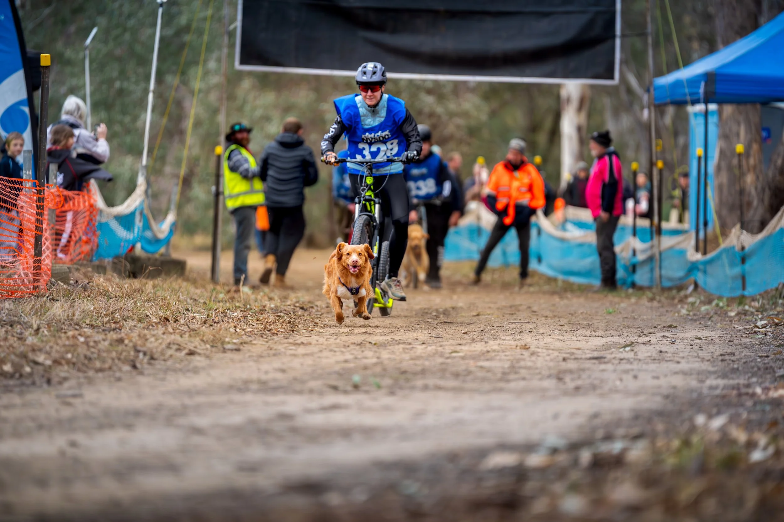 A person riding a bicycle with a dog running alongside on a dirt trail at a race event, with spectators and volunteers in the background.