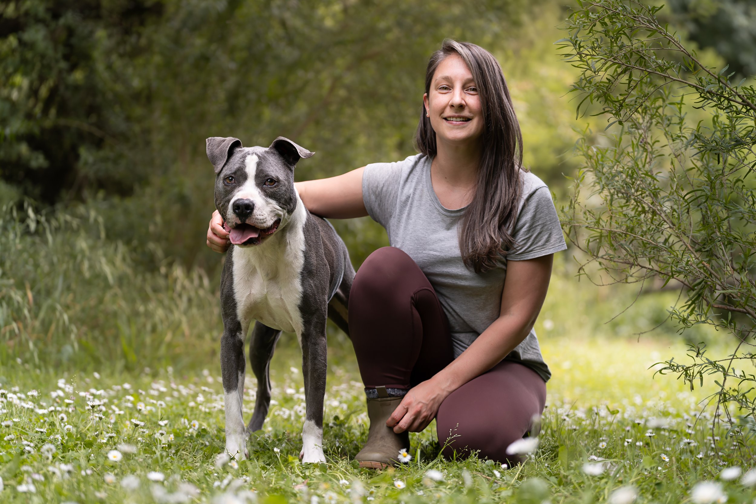 A woman kneeling on grass with small white flowers, smiling, with her arm around a gray and white dog, in a green outdoor setting.