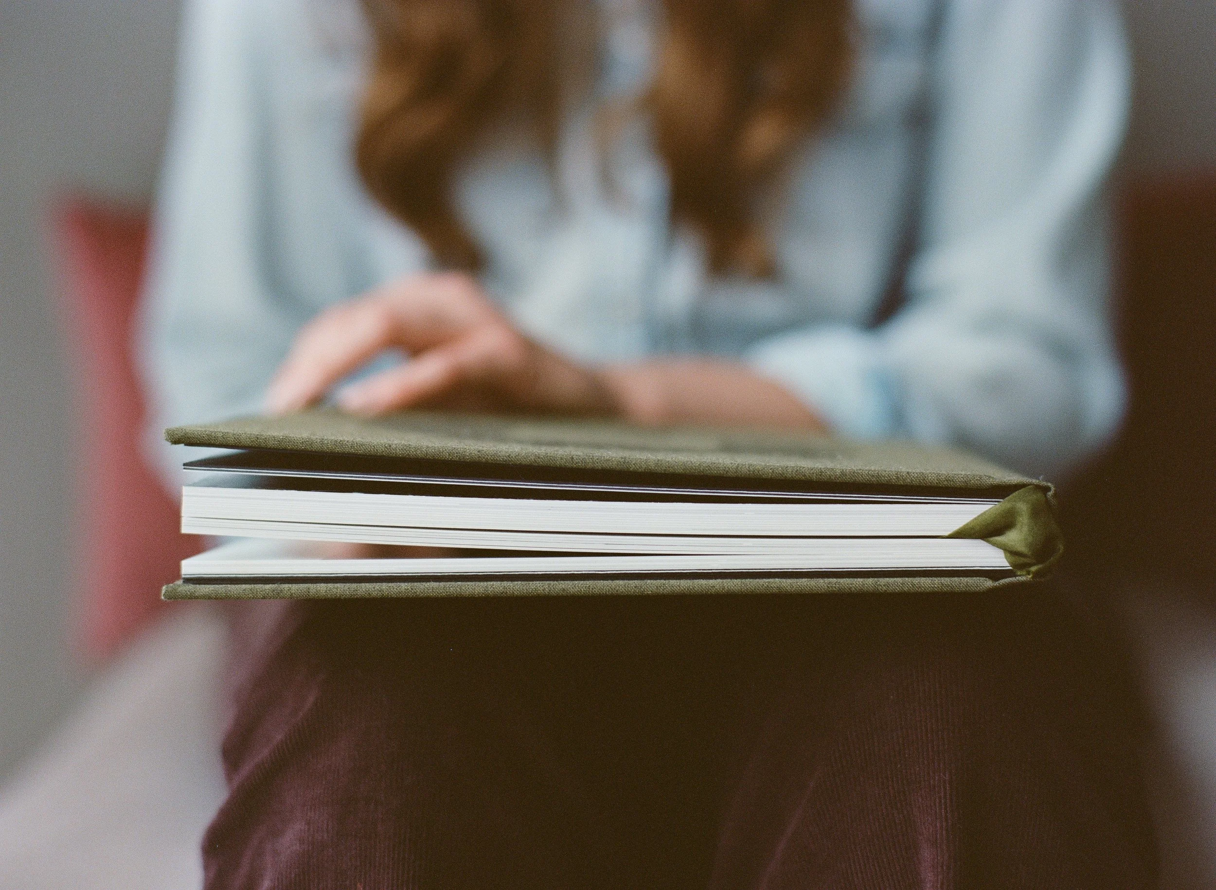 A person sitting with a green notebook open on their lap, holding a pen, with long, curly hair and wearing a light-colored top.