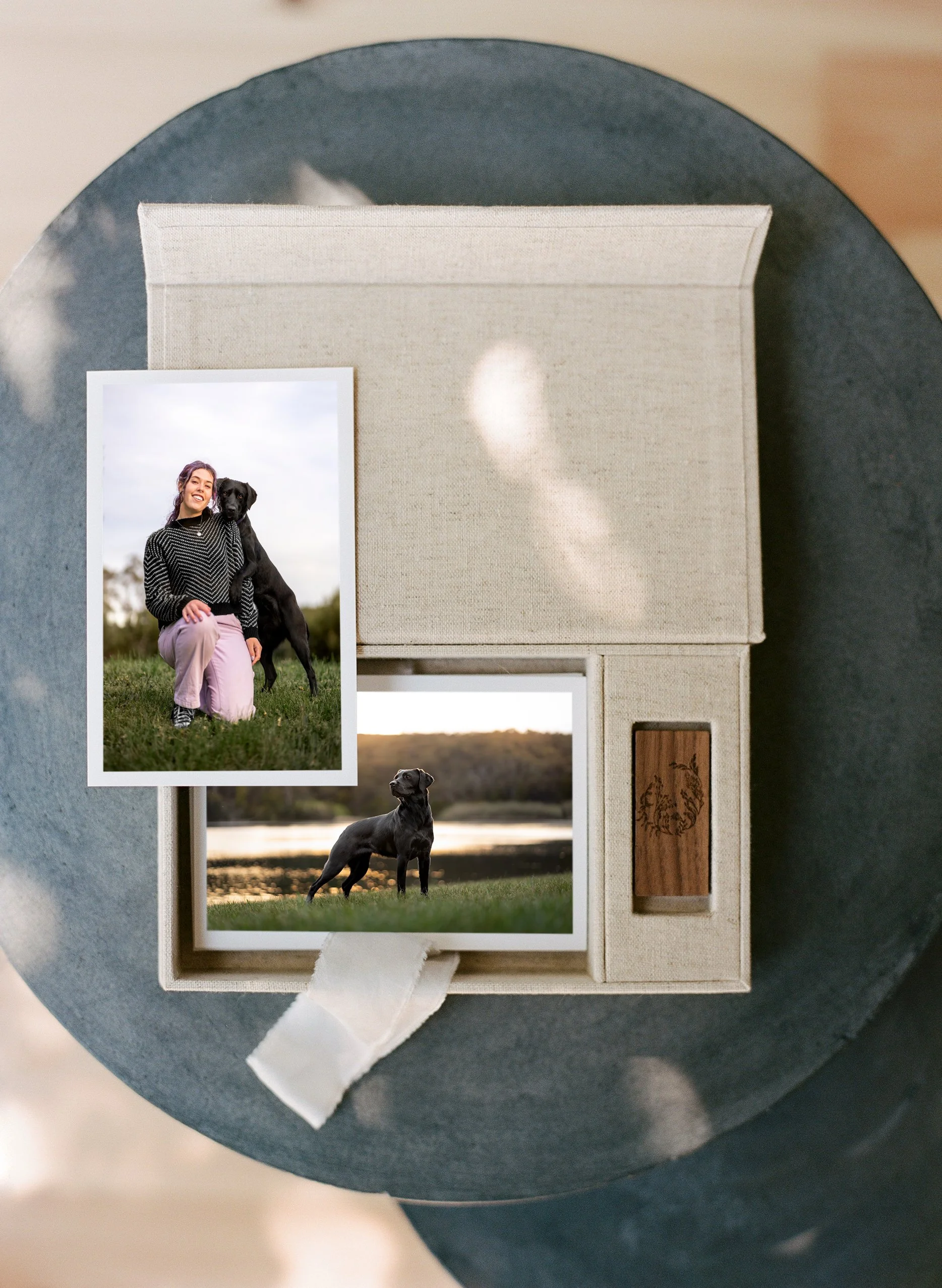 A box with two printed photos of a woman and a black dog, placed on a round dark surface with blurred light spots in the background.