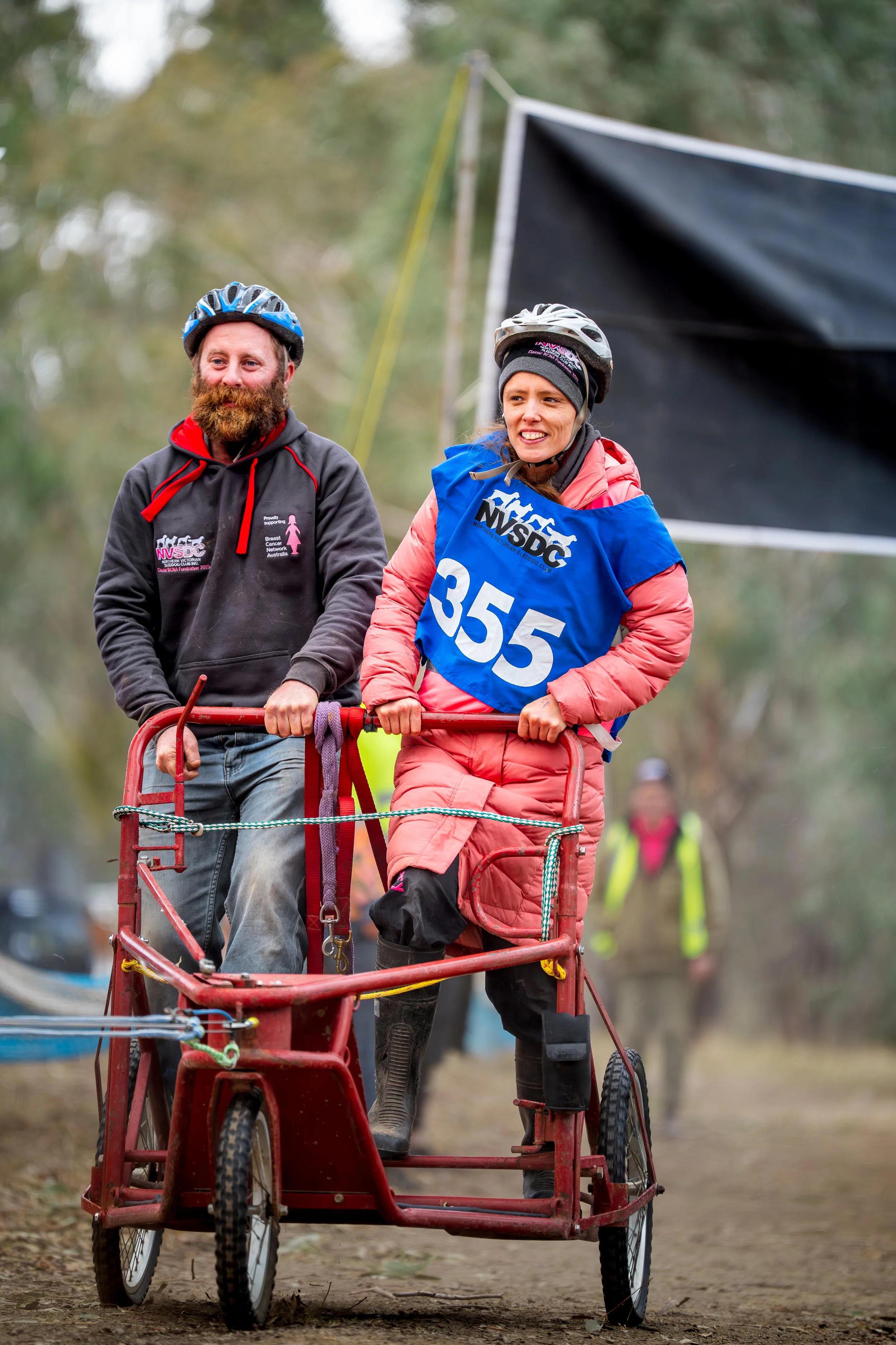 Two cyclists on a velomobile, one woman and one man, outdoors on a dirt trail with trees in the background. The woman is wearing a pink winter jacket, black knee-high boots, a gray hat, and a blue vest with the number 355 and the NSDC logo. The man i