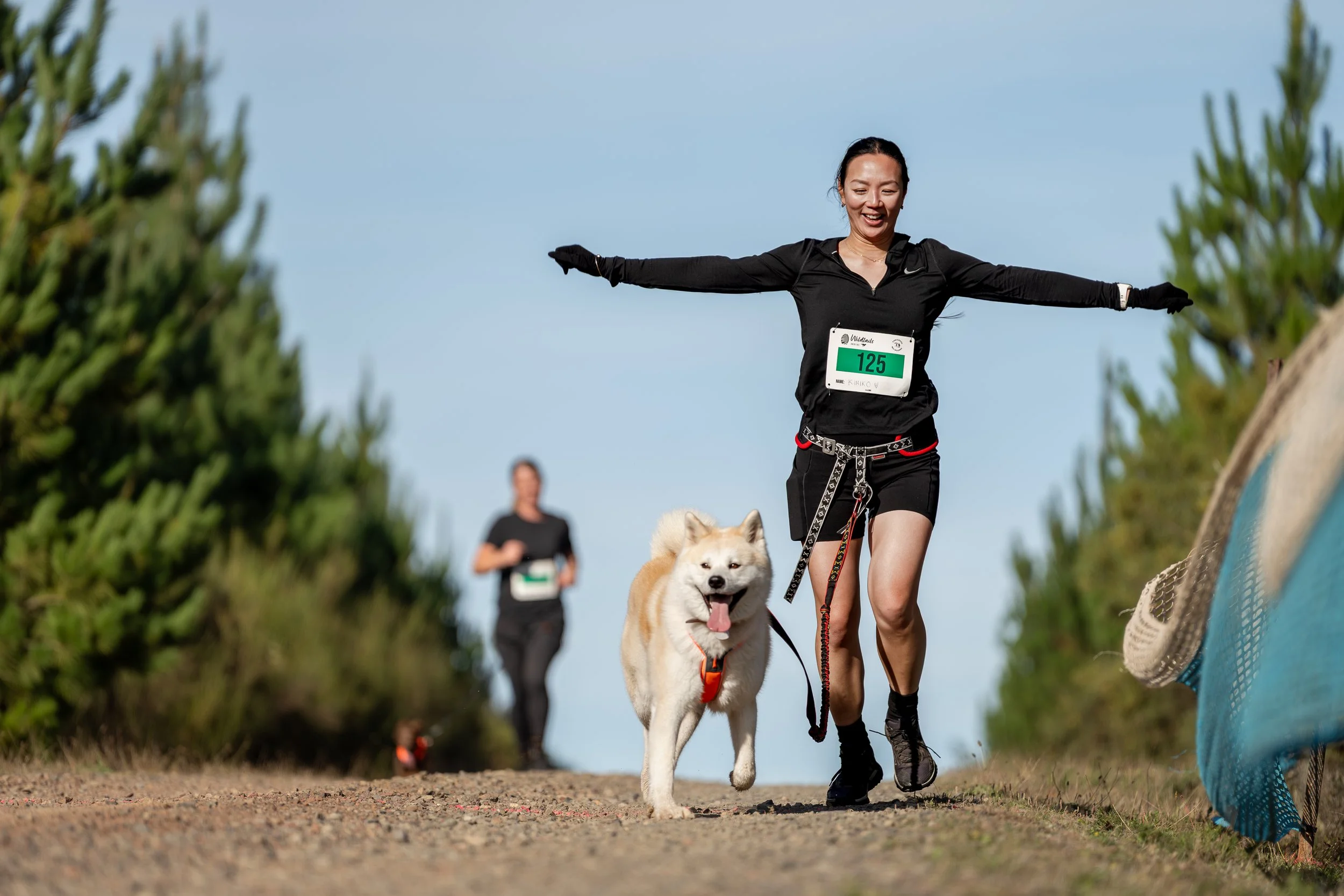 A woman running in a race with a dog on a leash, with another woman running behind them. The woman is wearing black athletic wear with a race bib number 125, and the dog appears happy with its tongue out. The race takes place outdoors on a trail with