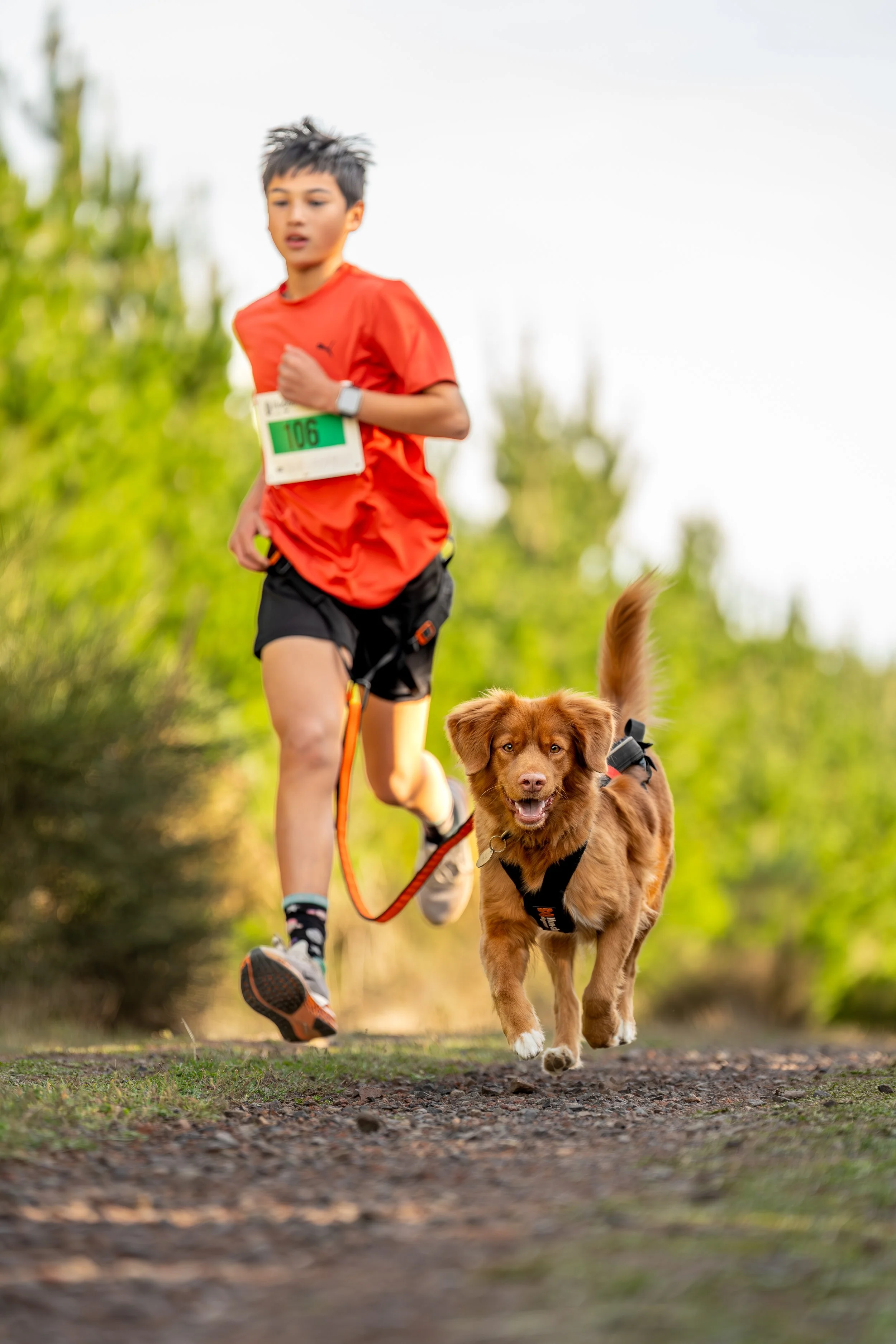 A boy running with a brown dog on a trail in a forested area.