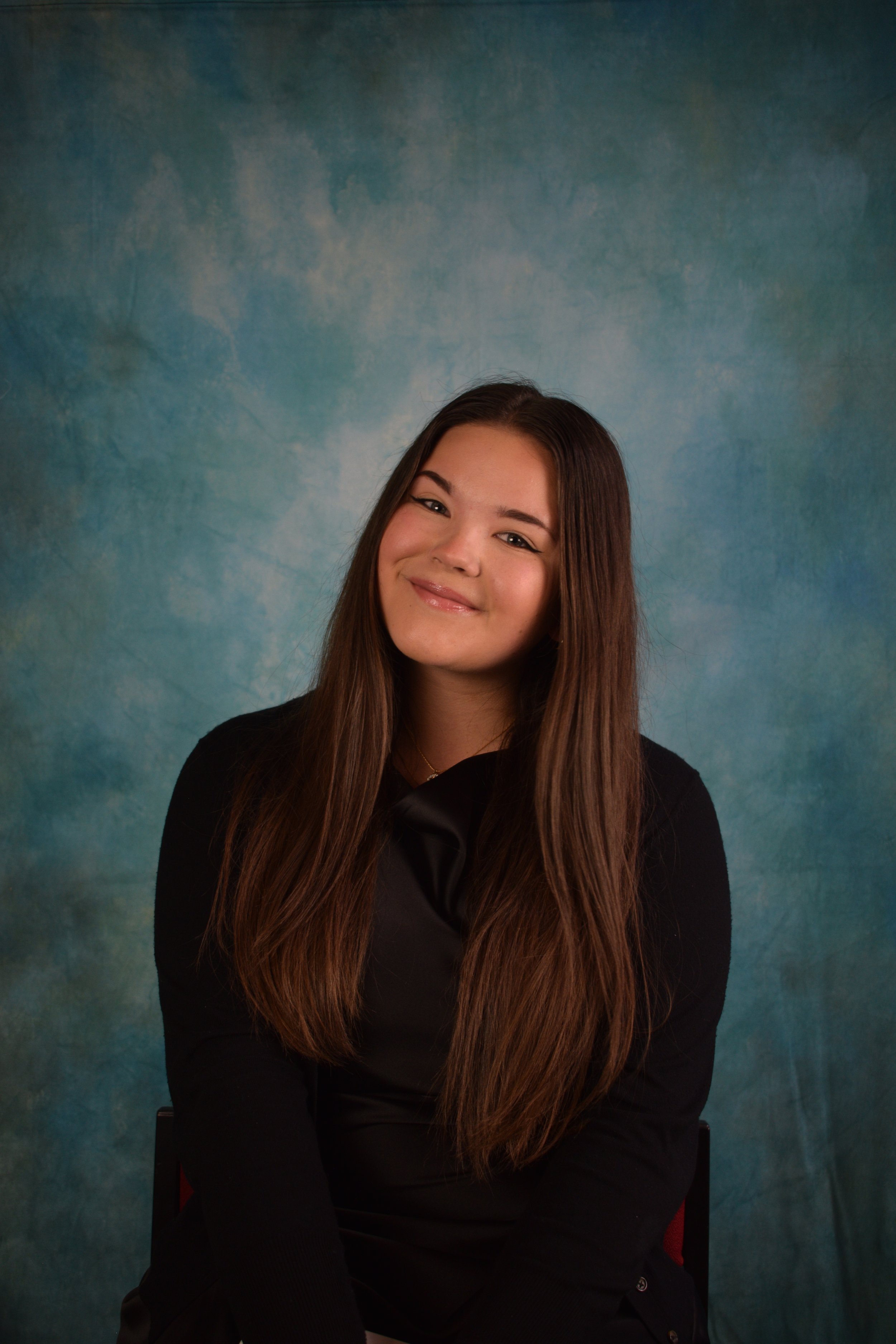 Holly is a young woman with long brown hair, smiling while sitting against a blue textured background, wearing a black top.