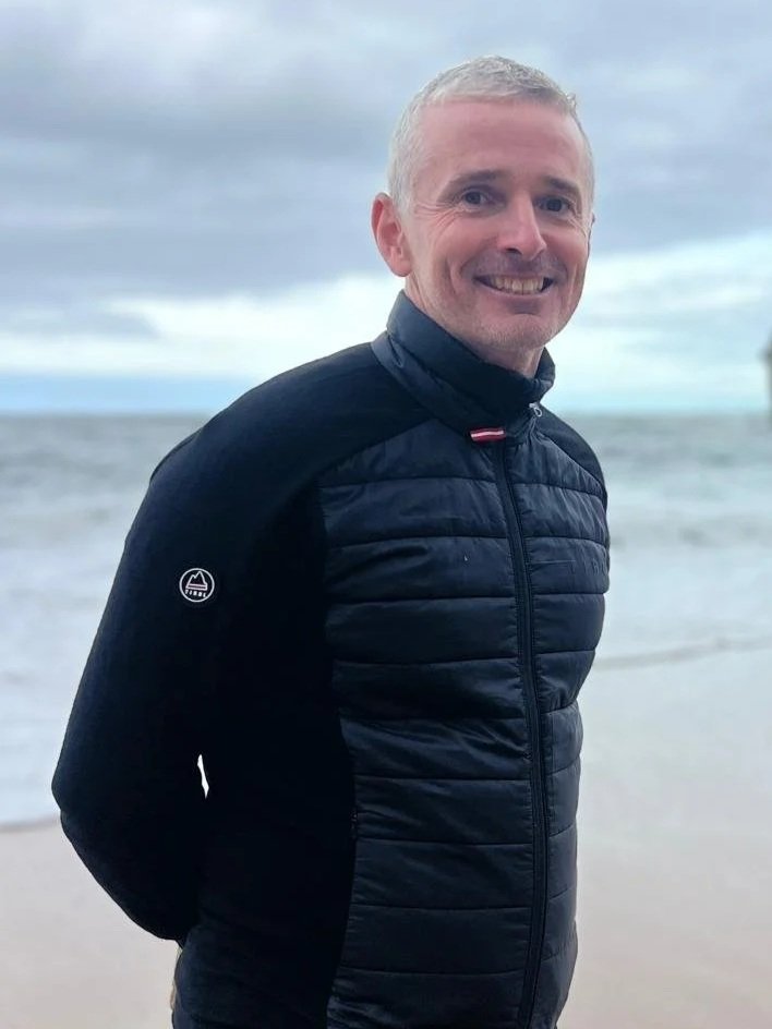 Smiling man in a black jacket standing on a beach with ocean and cloudy sky in the background.