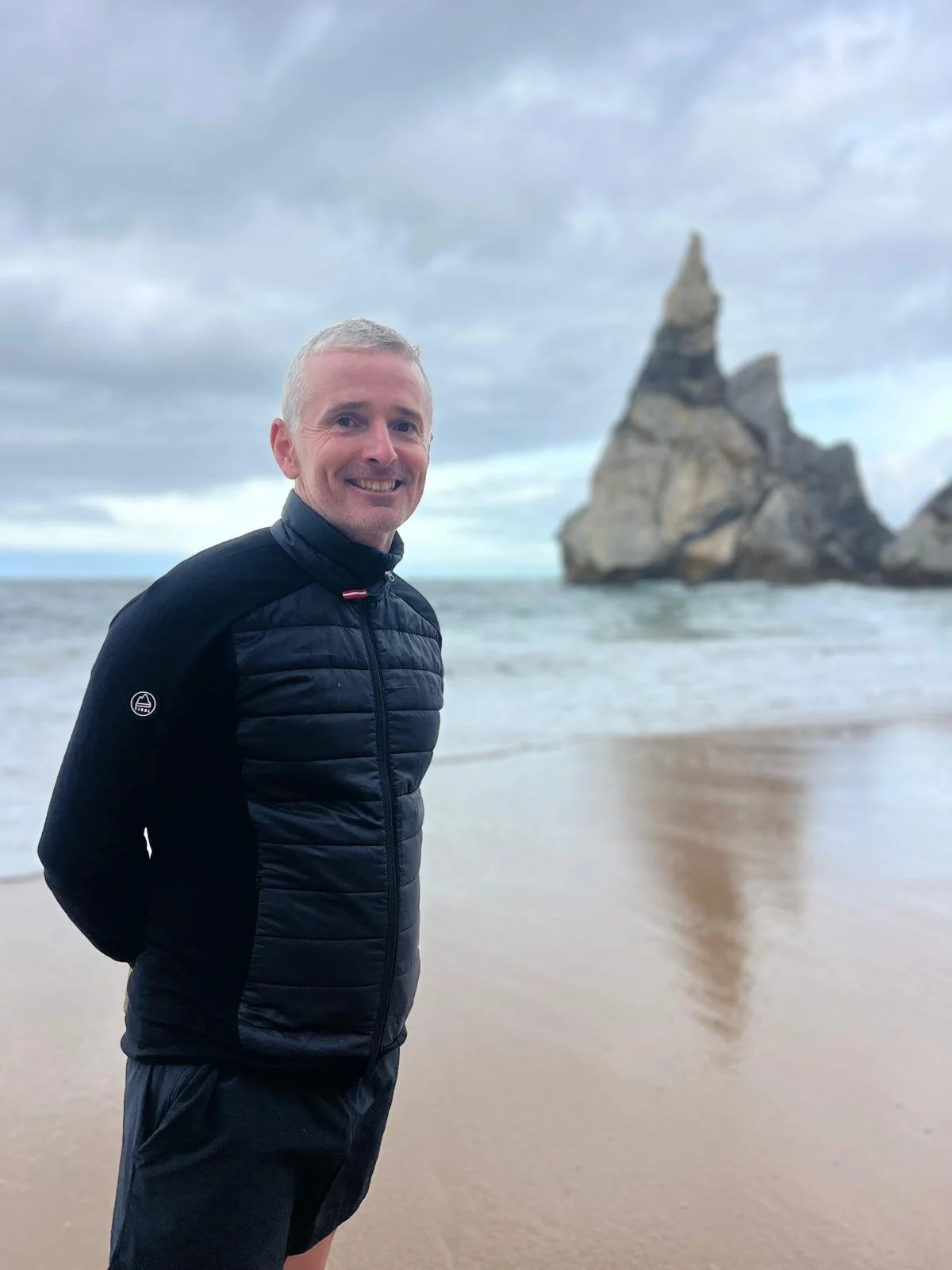 A man in a black jacket stands on a beach with ocean waves and a large rocky formation in the background under a cloudy sky.