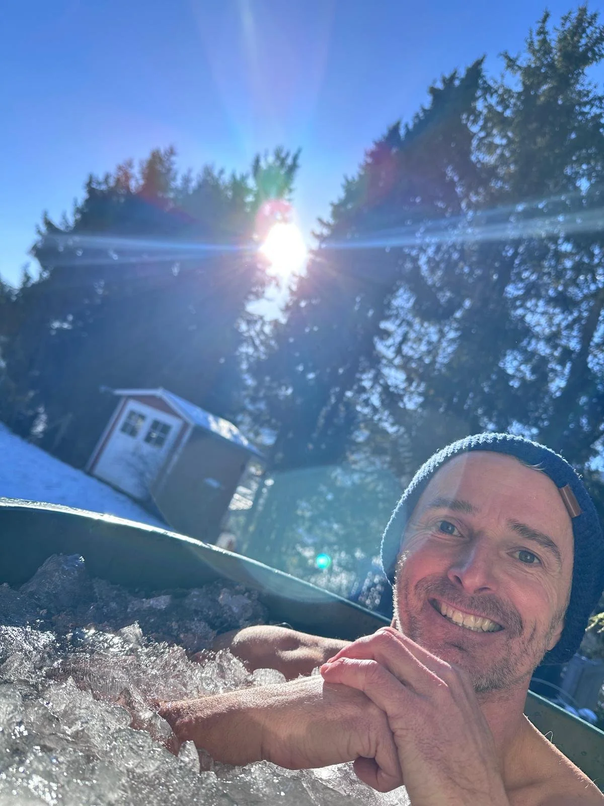 A smiling man relaxing in an outdoor hot tub with snow on his shoulders and a bright sun in the clear blue sky, surrounded by trees and a small shed in the background.
