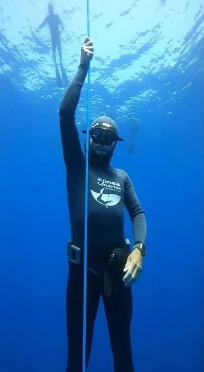 A scuba diver underwater holding a measuring stick, with another diver visible in the background.
