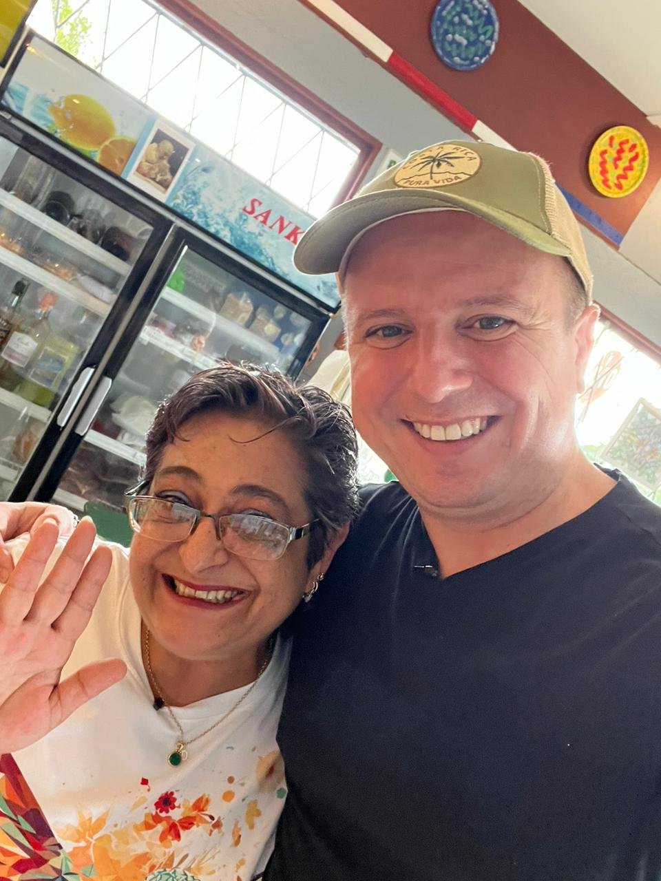 A smiling woman with glasses and short dark hair waving next to a smiling man wearing a beige hat. They are indoors, in front of a refrigerator with beverages and a top sign that reads 'SANK.' The woman is wearing a white shirt with colorful floral patterns, and the man is in a black shirt.