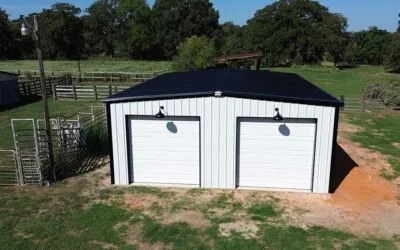 A white metal garage with two doors and black roof, situated on a grassy area with trees and fenced paddocks in the background.