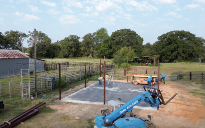 Construction site with a concrete foundation and blue crane equipment, surrounded by fencing and trees in the background.