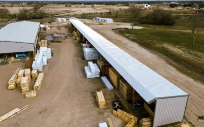 Aerial view of a storage or warehouse facility with stacked pallets and boxes under a long metal roof, surrounded by open land.