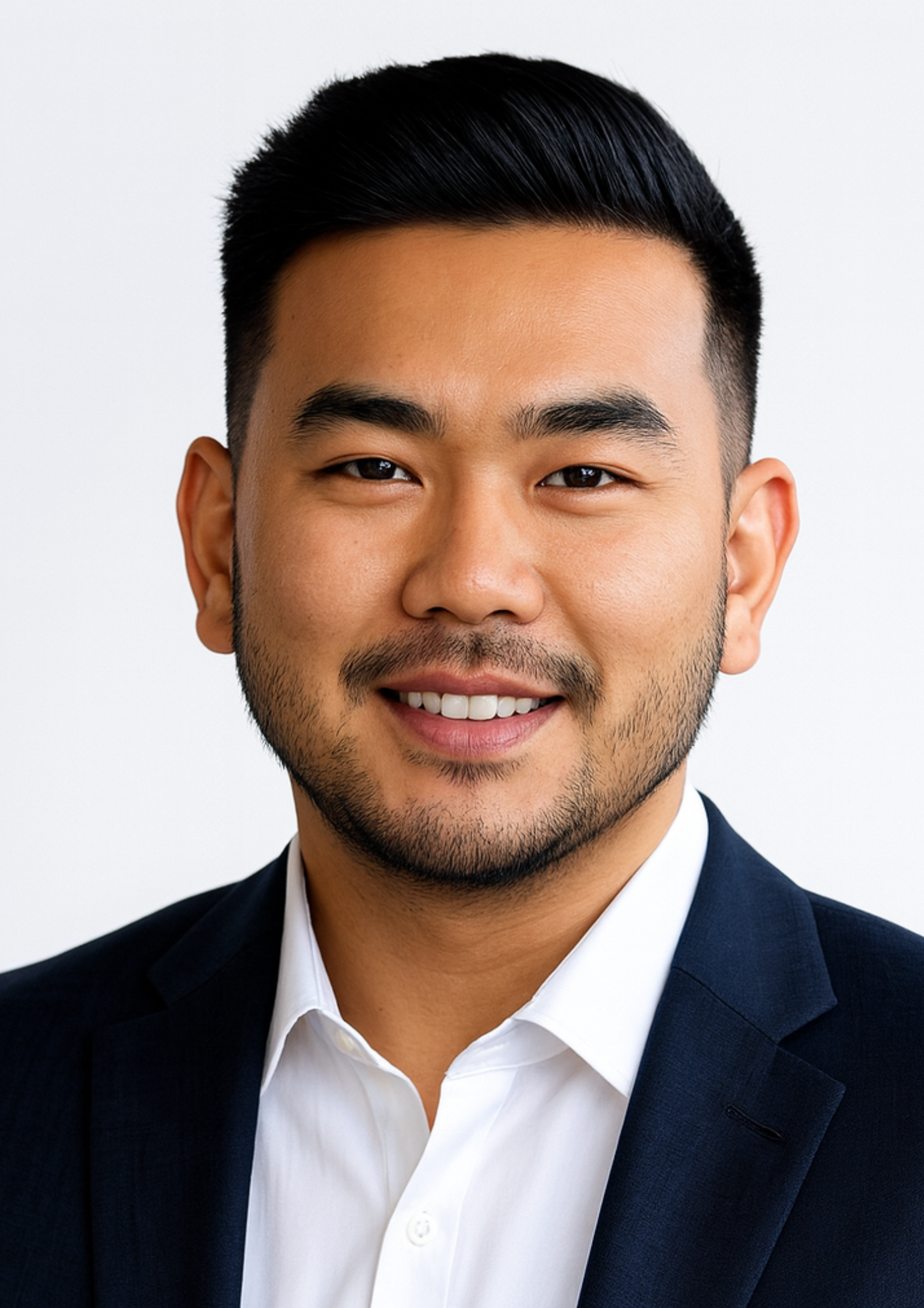 A professional headshot of a man with short black hair, a beard, wearing a dark suit jacket and a white dress shirt, smiling against a plain light background.