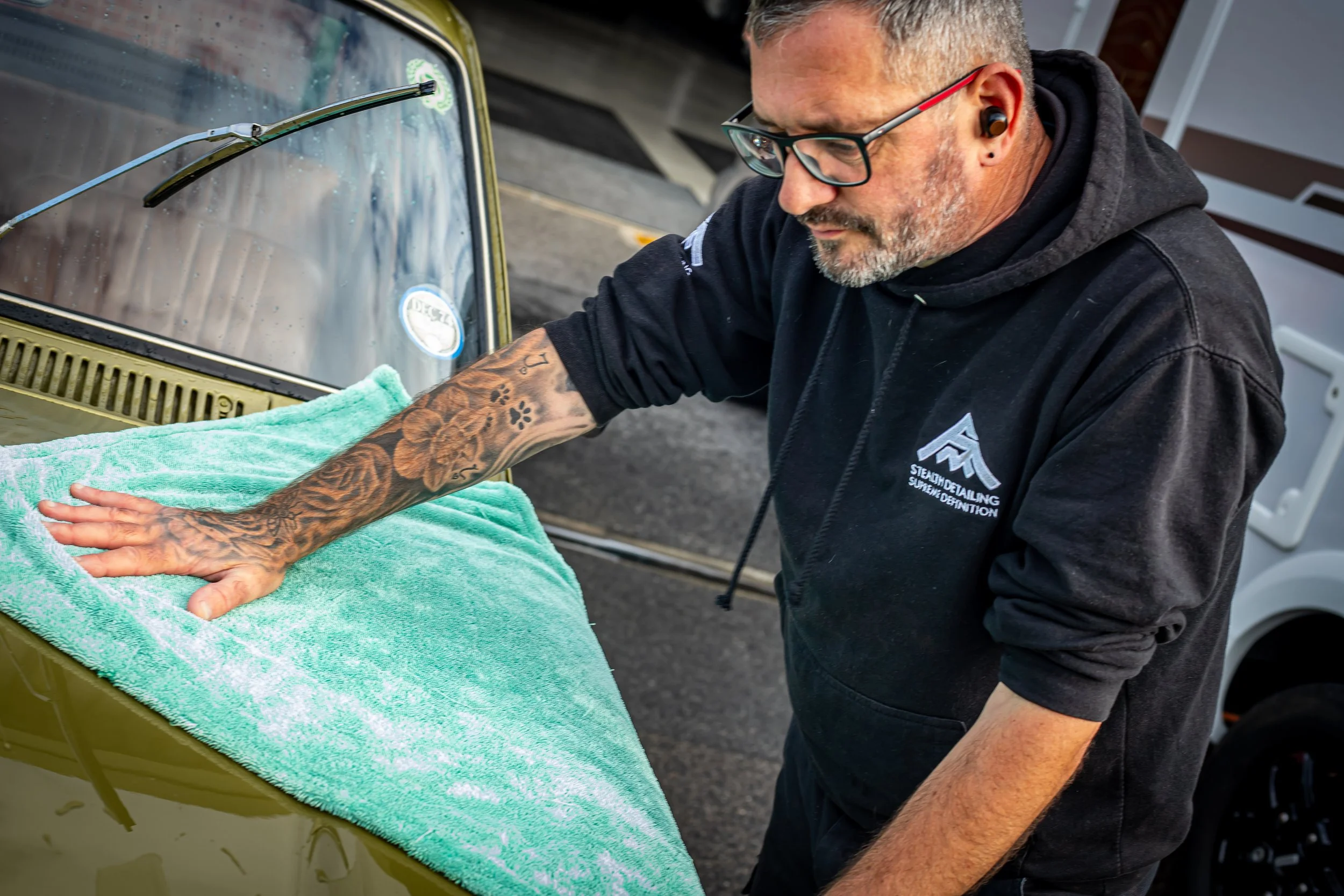 A man with tattoos on his arm wearing glasses and a black hoodie, labeled 'Stealth Detailing Supreme Detailing,' is polishing or cleaning a yellow or green vintage car with a green towel. He is using earbuds and working outdoors.