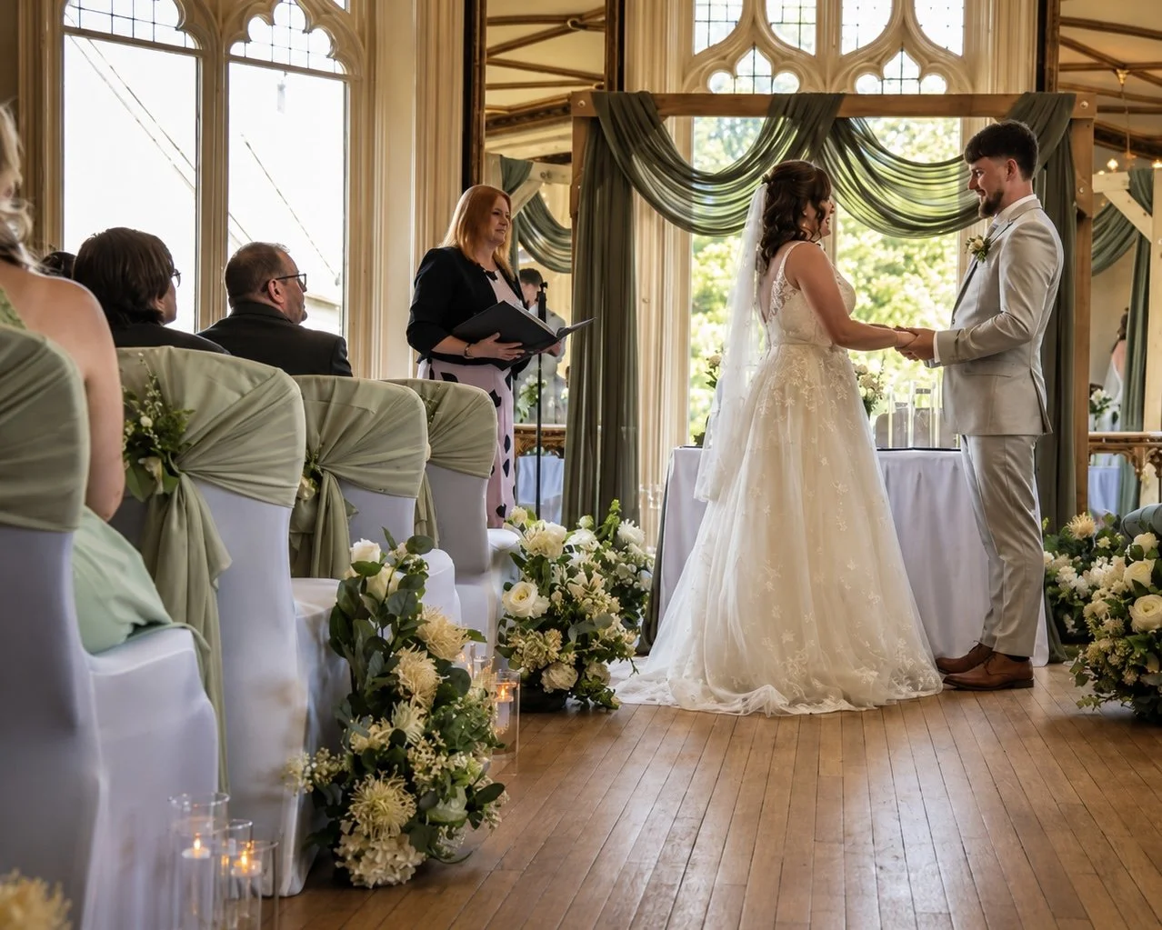 A bride and groom holding hands and exchanging vows during a wedding ceremony in a decorated indoor venue with large windows and floral arrangements.