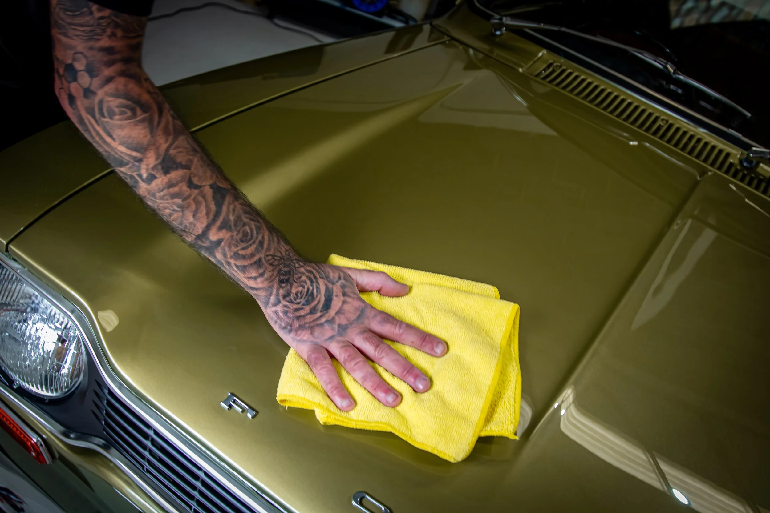 A person with a tattooed arm is cleaning the hood of a gold-colored vintage car with a yellow cloth.