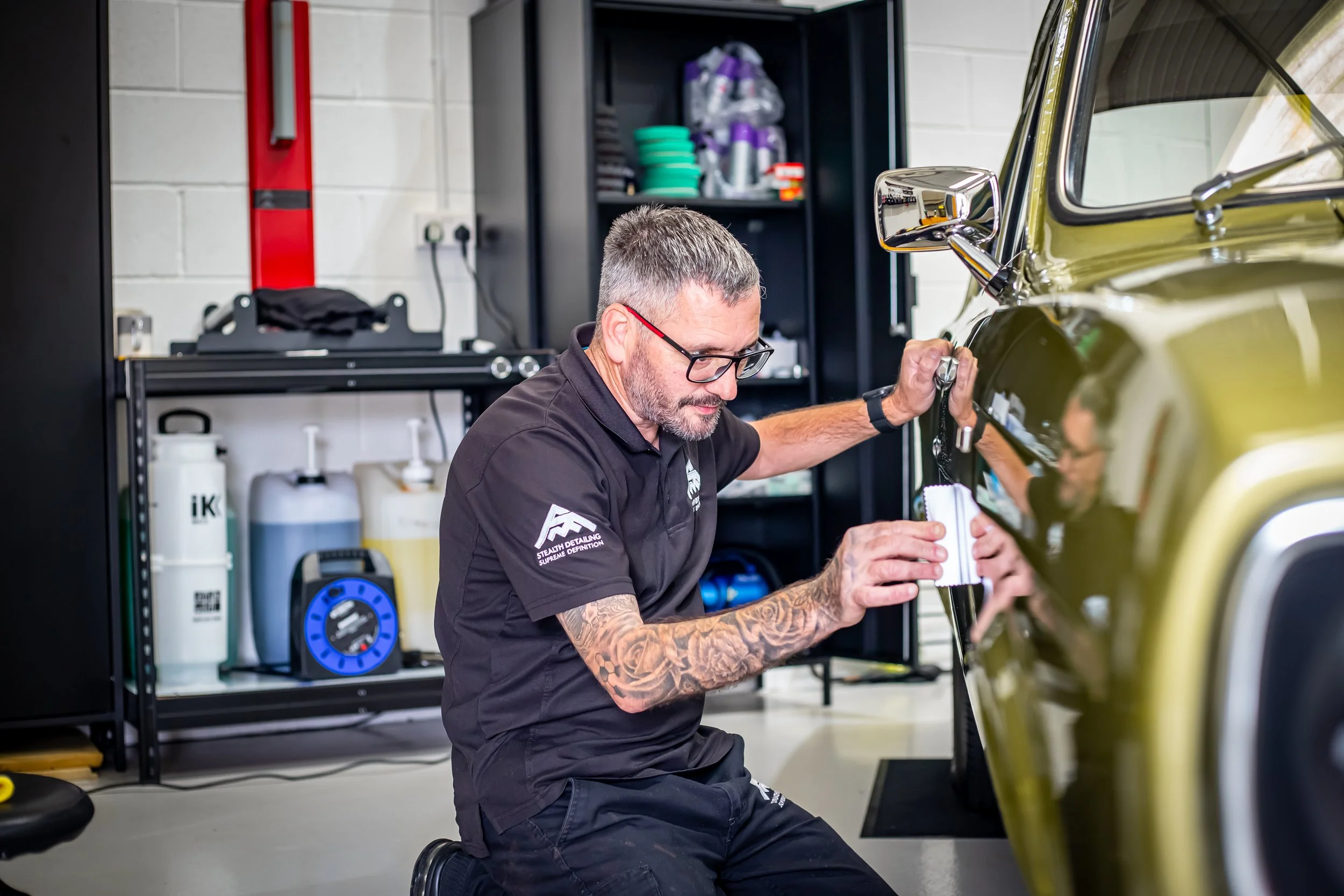 A man with tattoos, short hair, and glasses kneeling and polishing a green classic car in a garage.