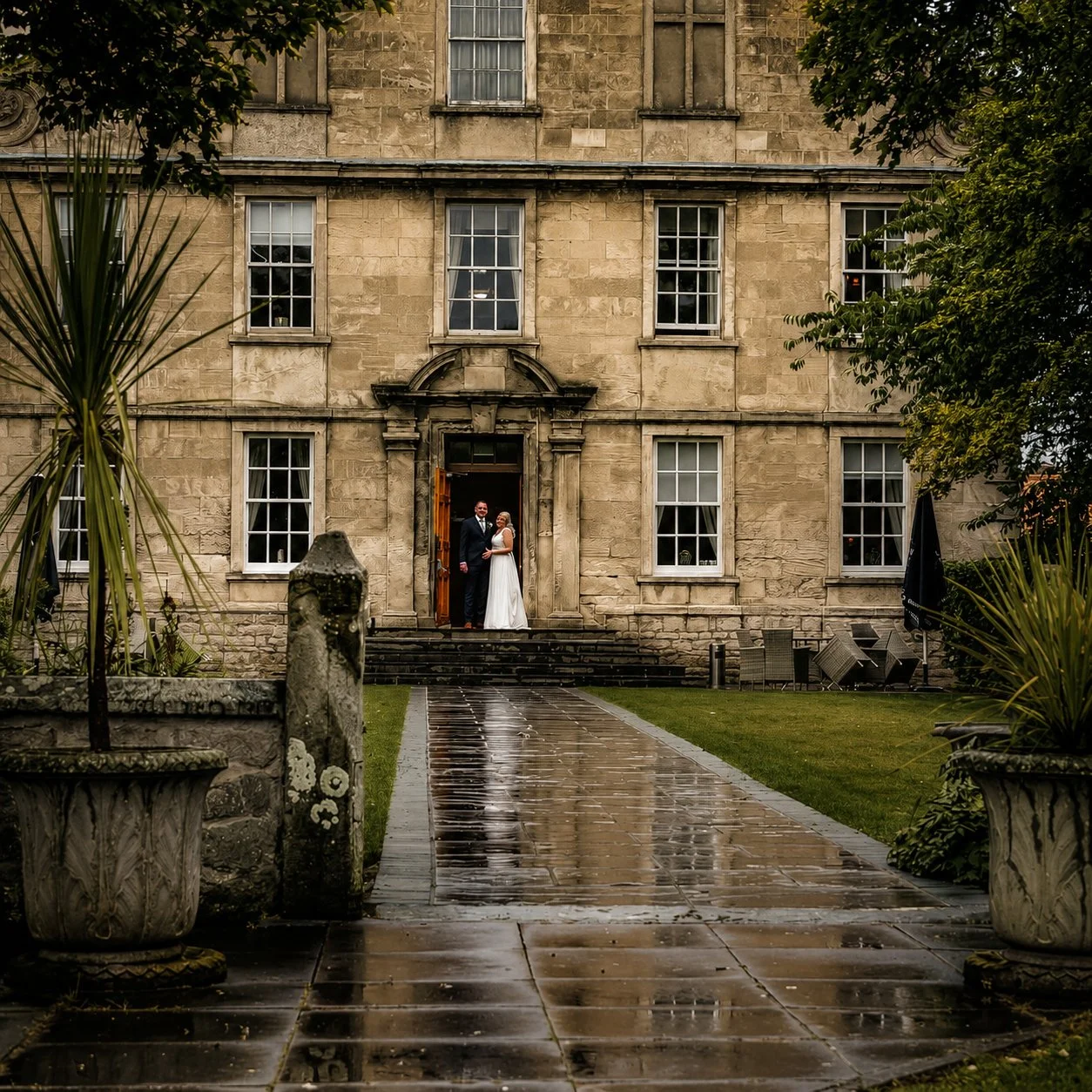 A bride and groom standing on the steps of a historic stone building with large windows, surrounded by greenery, on a rainy day.
