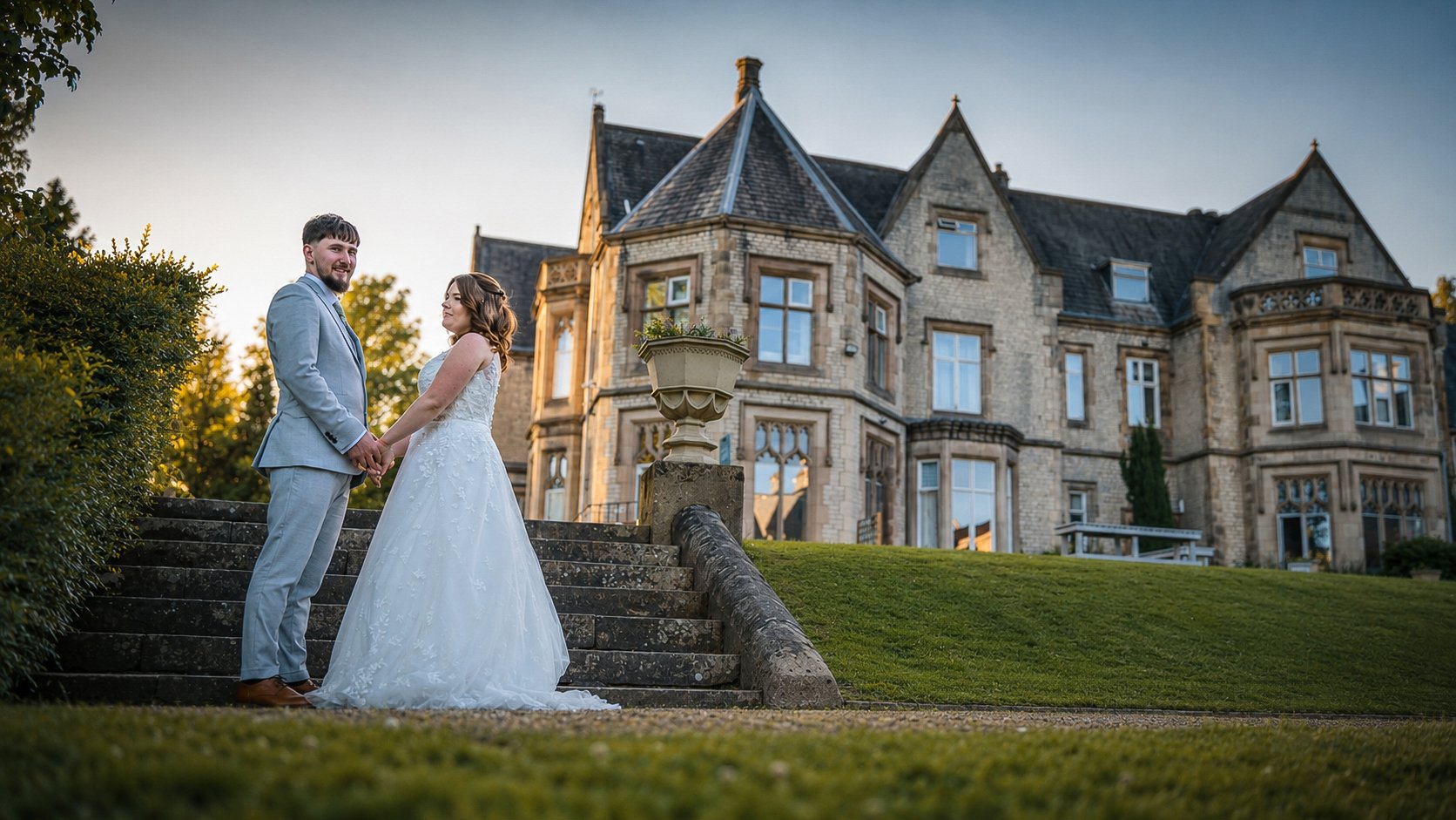 A bride and groom holding hands and looking at each other on stone steps outside a large historic mansion during sunset.
