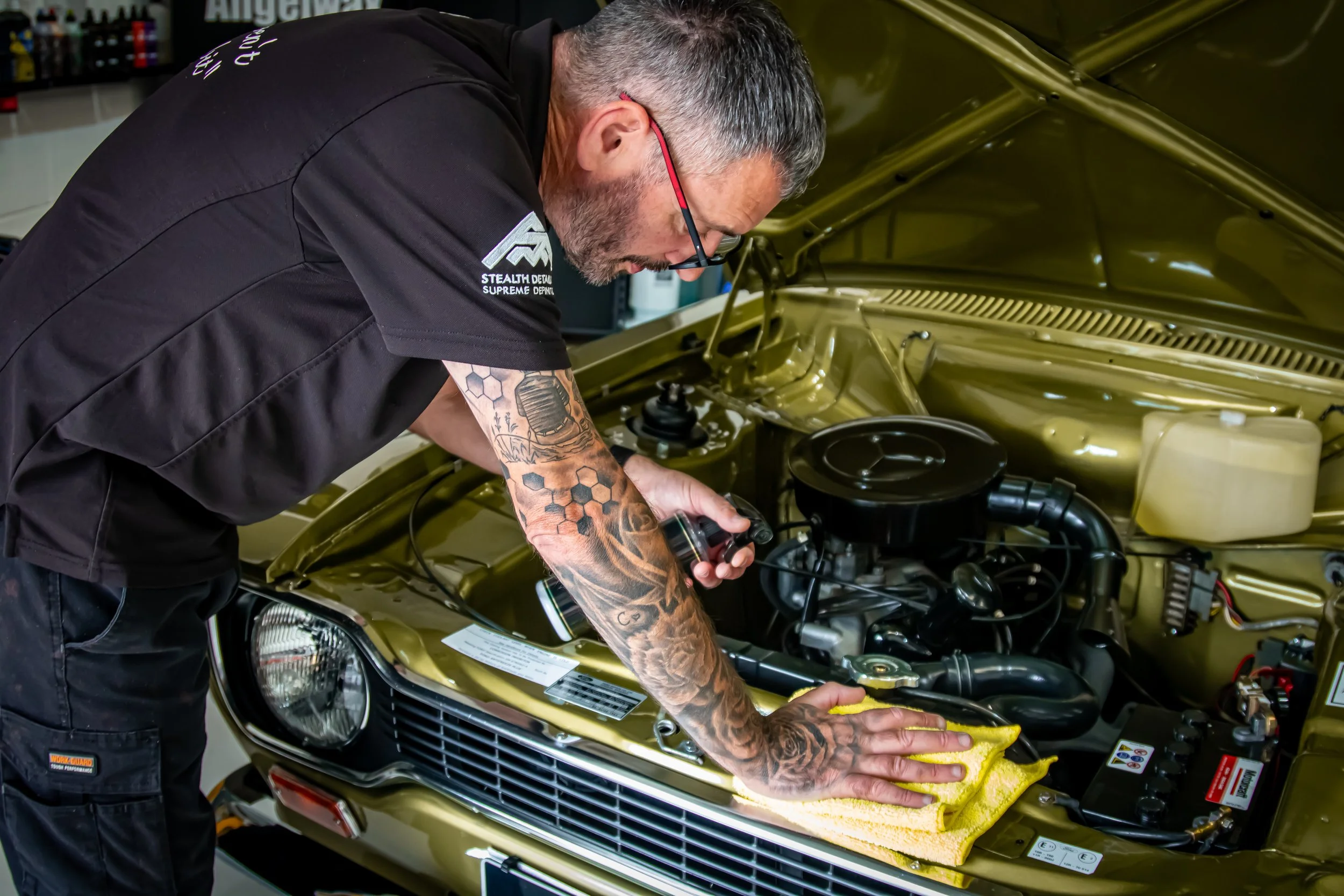 A man with tattoos and glasses inspecting or working on the engine of a gold-colored car in a garage or auto shop.